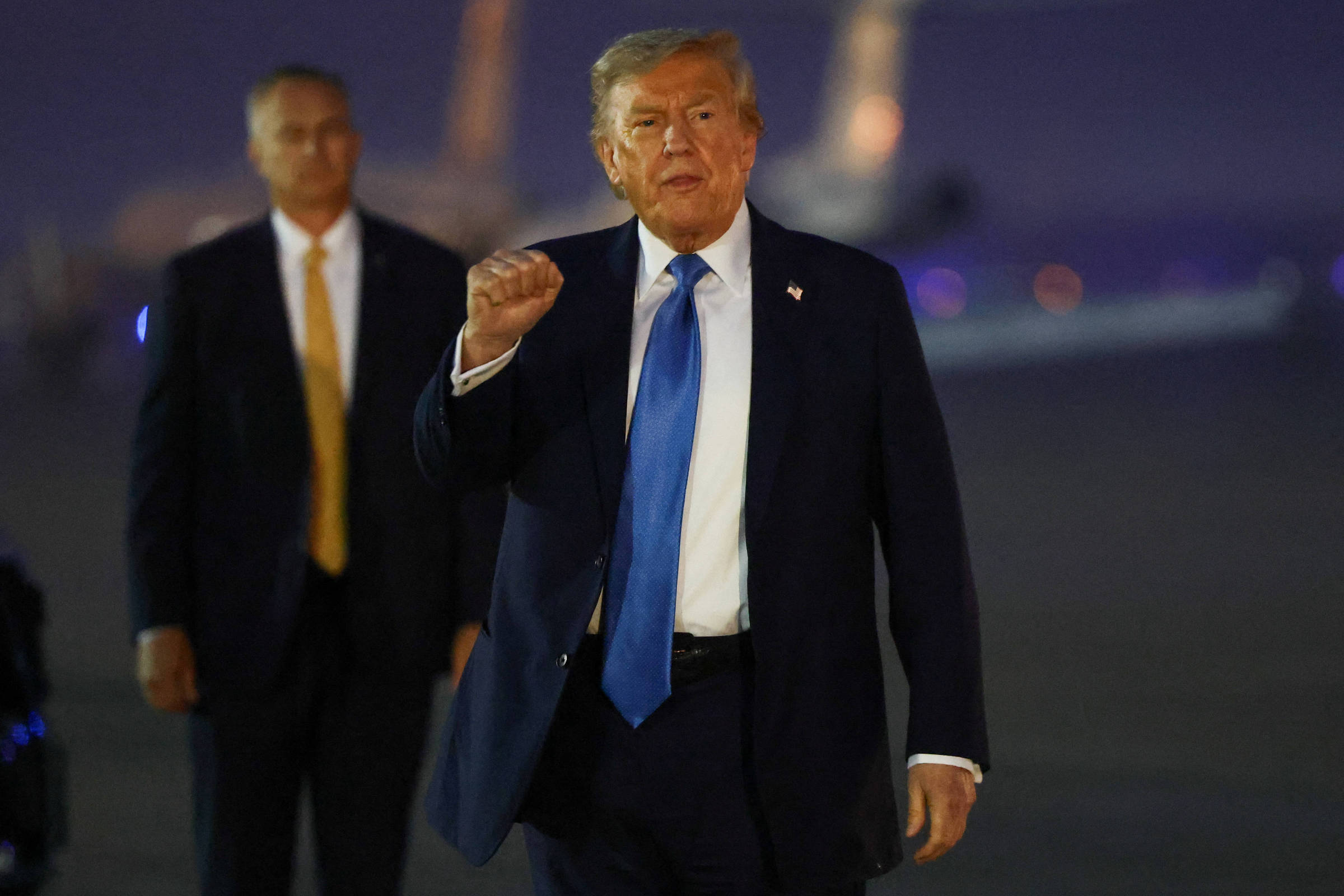 Imagem mostra Donald Trump. um homem vestido com um terno escuro e uma gravata azul e que caminha em direção à câmera, fazendo um gesto com a mão. Ao fundo, há um avião e outra pessoa em traje formal, que parece ser um segurança, observando. A cena ocorre em um ambiente noturno, com iluminação suave.