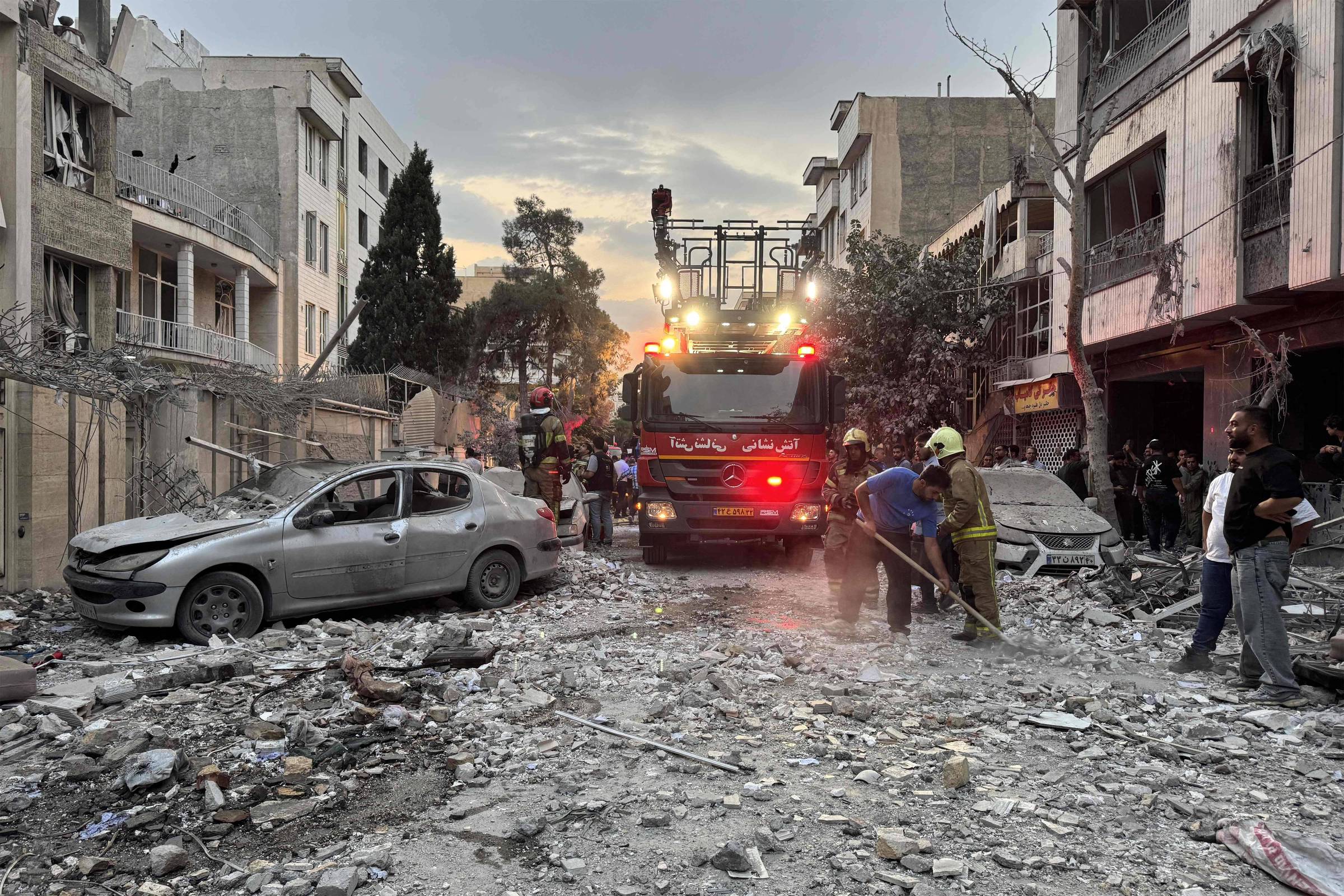 A imagem mostra uma cena de destruição em uma área urbana, com prédios danificados e escombros espalhados pelo chão. Um caminhão de bombeiros está presente, com luzes acesas, enquanto várias pessoas, incluindo bombeiros e civis, estão trabalhando para limpar os destroços. Há carros danificados na calçada e árvores ao fundo, com um céu nublado ao entardecer.