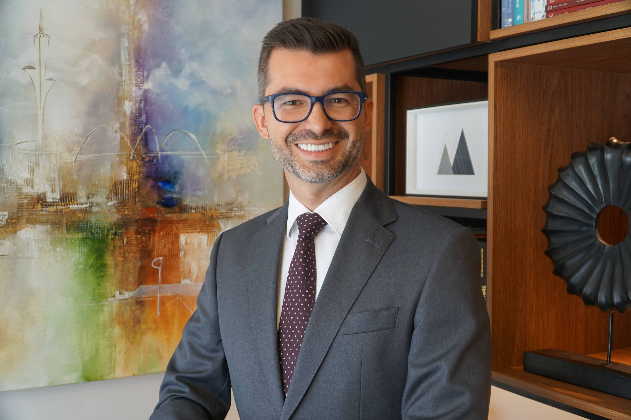 Um homem sorridente está posando em um ambiente moderno e bem iluminado. Ele usa um terno cinza e uma gravata com padrões. Ao fundo, há uma pintura abstrata com cores vibrantes e elementos decorativos como prateleiras com livros e objetos de arte. O homem tem cabelo curto e usa óculos.