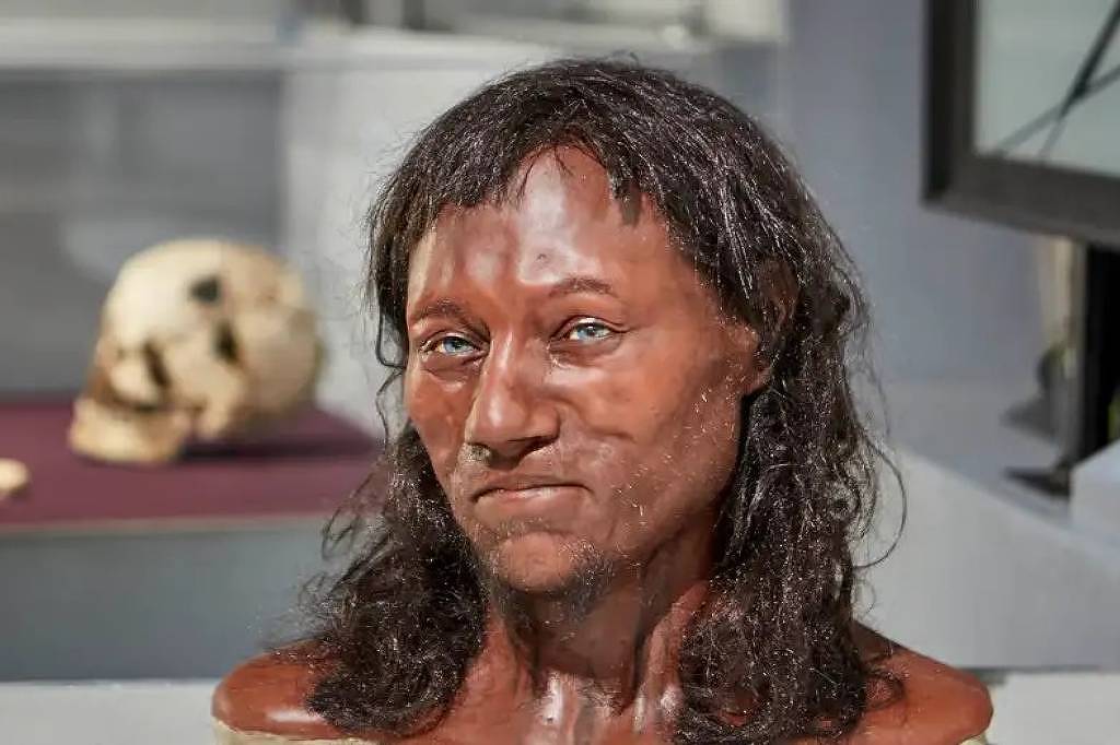 A imagem mostra uma reconstrução facial em 3D de um homem pré-histórico, com pele escura e cabelo escuro e liso. O busto é apresentado em um ambiente de museu, com um crânio e ossos visíveis ao fundo, sugerindo um contexto arqueológico. A expressão facial é neutra, e a reconstrução parece detalhada e realista.