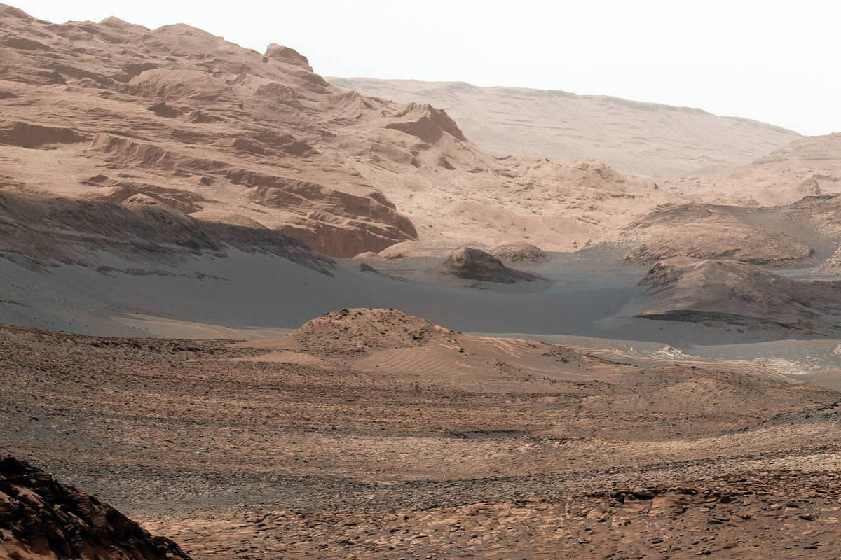 A imagem mostra uma vasta paisagem marciana, caracterizada por montanhas e vales áridos. O terreno é predominantemente rochoso e acinzentado, com formações geológicas visíveis ao fundo.