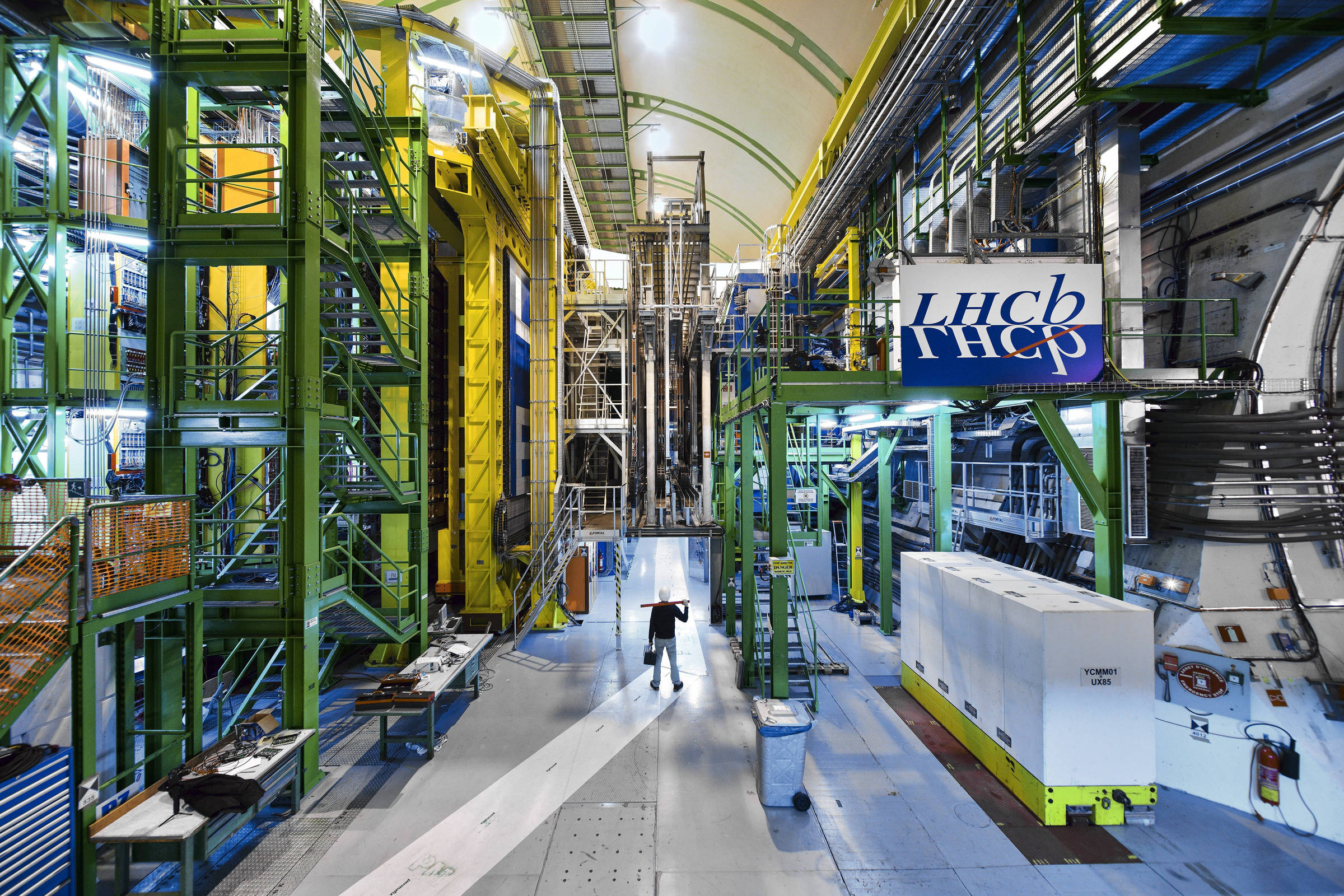 A imagem mostra o interior do experimento LHCb, localizado no Grande Colisor de Hádrons (LHC). O ambiente é amplo e industrial, com estruturas metálicas verdes e amarelas, além de equipamentos científicos. No centro, há uma pessoa em pé, que parece estar observando ou operando algum equipamento. O chão é de concreto e há diversos cabos e tubos visíveis nas paredes.