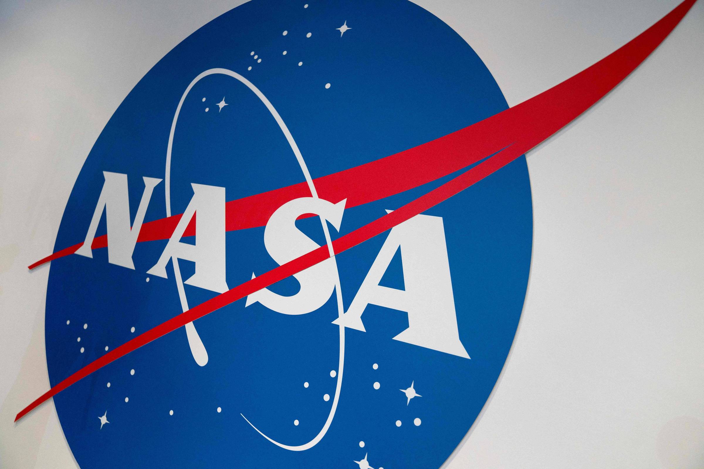 A imagem mostra o logotipo da NASA, que consiste em um círculo azul com estrelas brancas ao fundo. Dentro do círculo, as letras