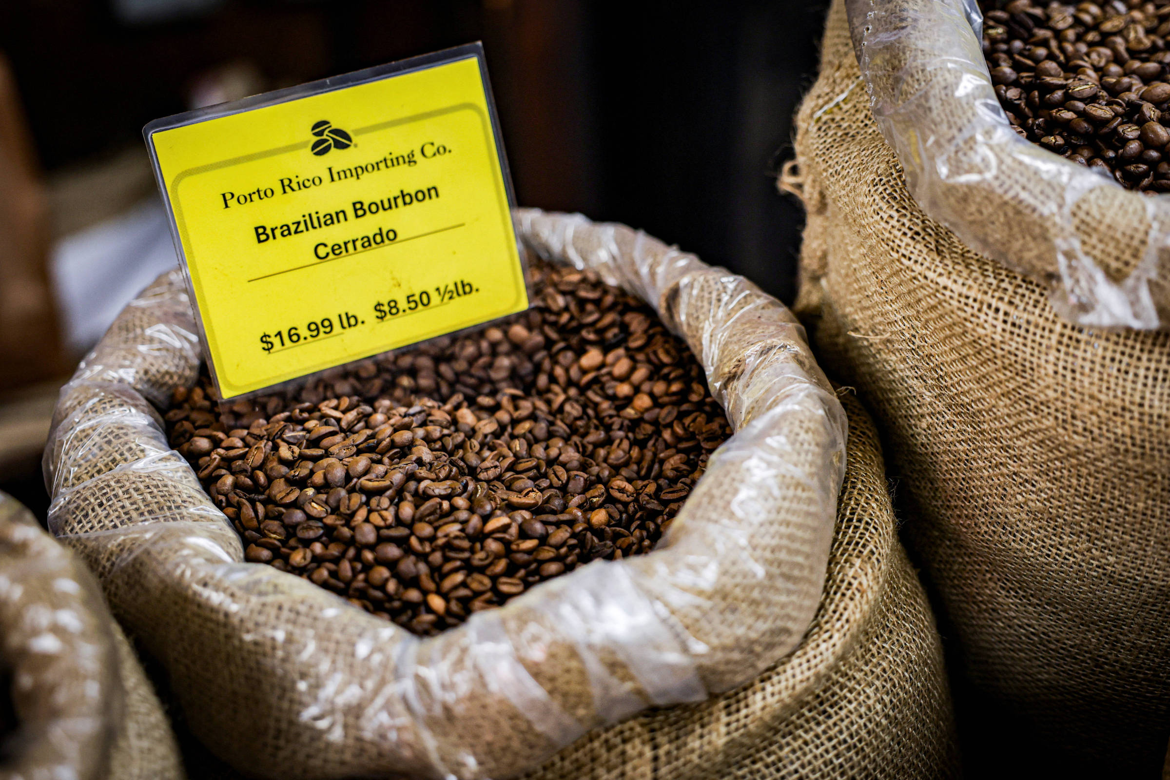 A imagem mostra sacos de café com grãos de café torrados visíveis. Um dos sacos tem uma placa amarela com o texto