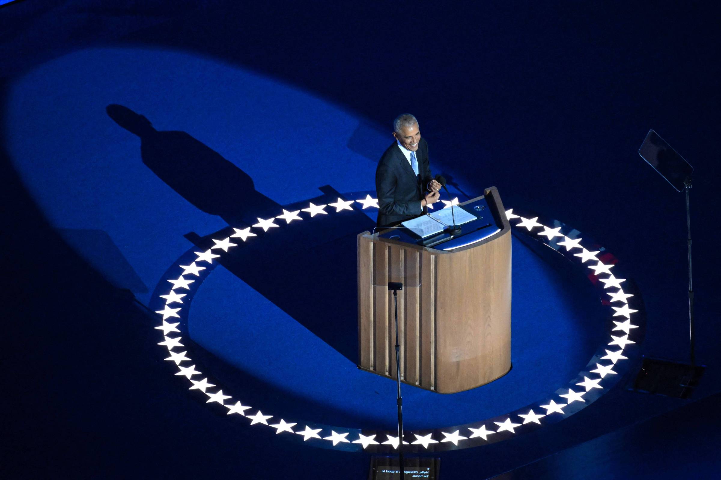 Um homem está em um palco, falando em um microfone, em frente a um púlpito de madeira. O fundo é iluminado em azul, com uma sombra projetada no chão. Ao redor do púlpito, há um círculo de estrelas brancas, que contrasta com o fundo azul. O homem parece estar gesticulando enquanto fala.