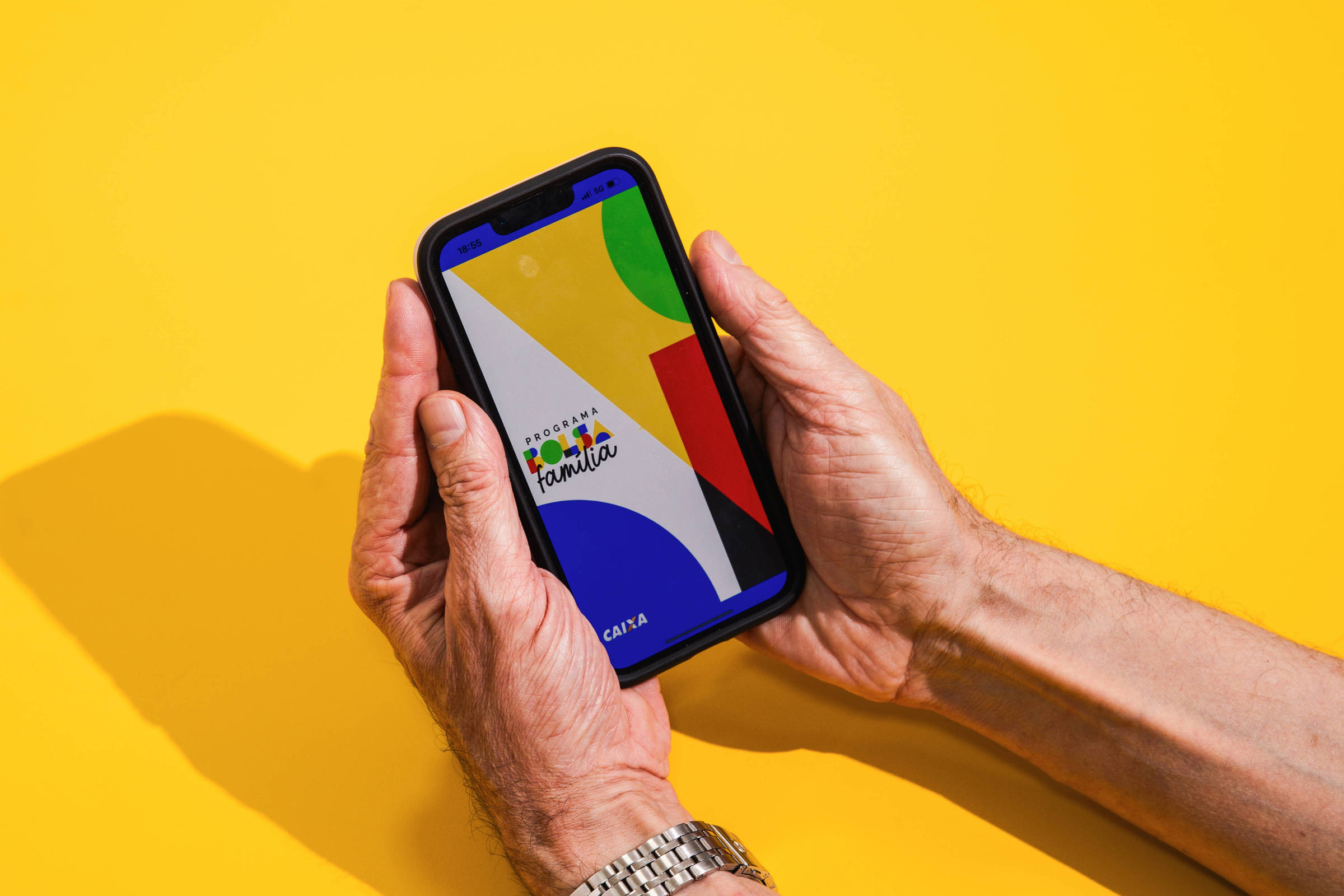 A imagem mostra uma mão segurando um celular. A tela do celular exibe um design colorido com formas geométricas em verde, vermelho, azul e amarelo, além do texto