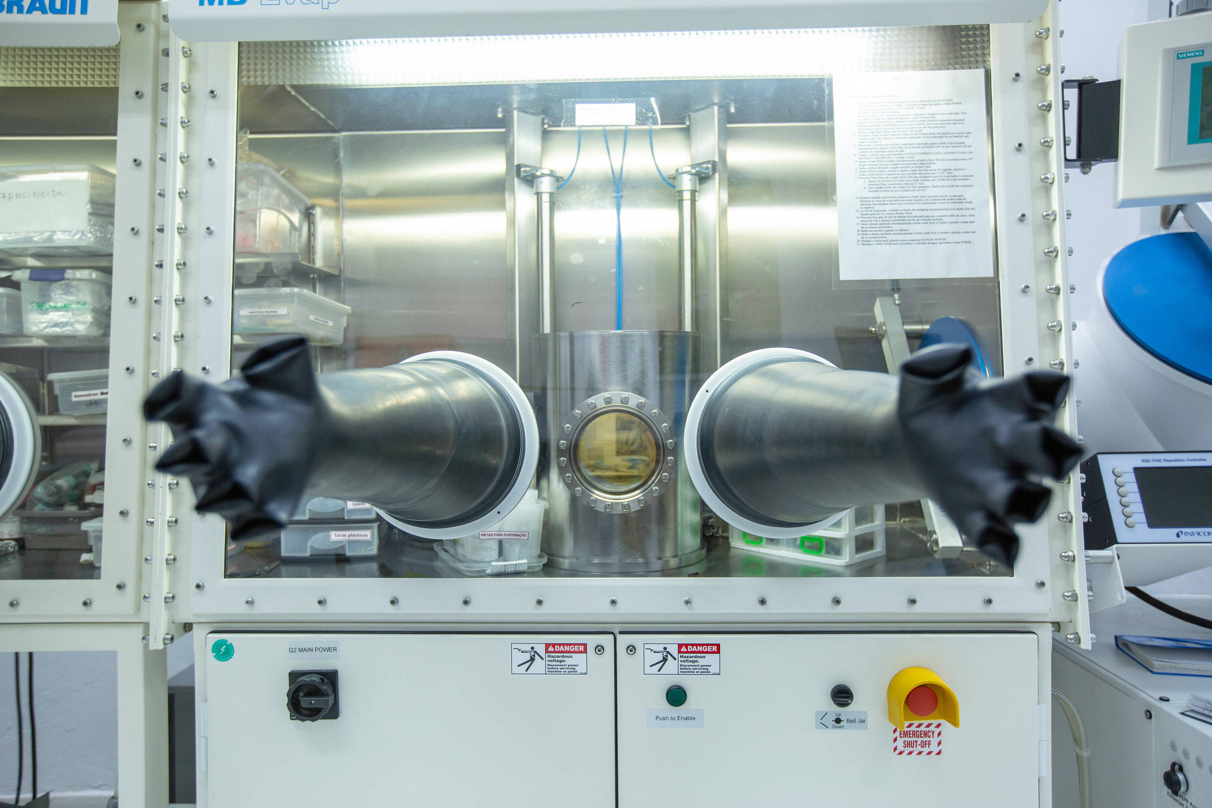 A imagem mostra um equipamento de laboratório com uma estrutura de vidro e metal. Na parte frontal, há duas luvas pretas saindo de aberturas circulares, permitindo a manipulação de materiais dentro do compartimento. O interior do equipamento é iluminado e contém um cilindro metálico central. Há também um painel de controle na parte inferior com botões e indicadores.