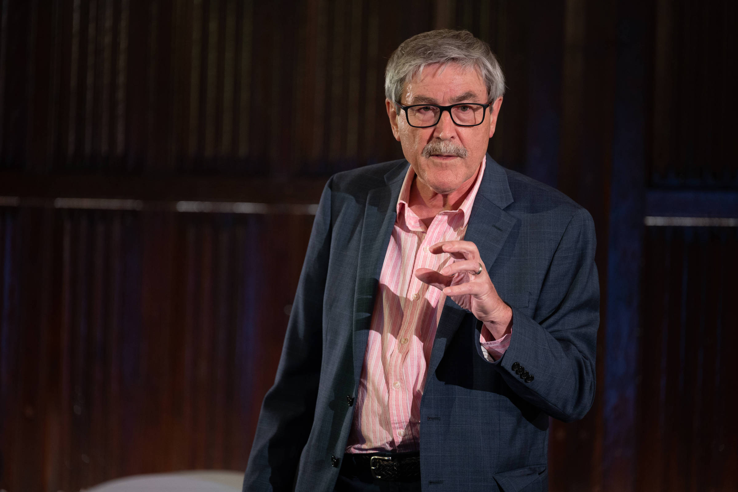 Um homem de cabelos grisalhos e óculos, vestido com um paletó escuro e uma camisa rosa, está em pé em um palco. Ele parece estar falando ou apresentando algo, gesticulando com a mão direita. O fundo é de madeira escura, sugerindo um ambiente de conferência ou palestra.