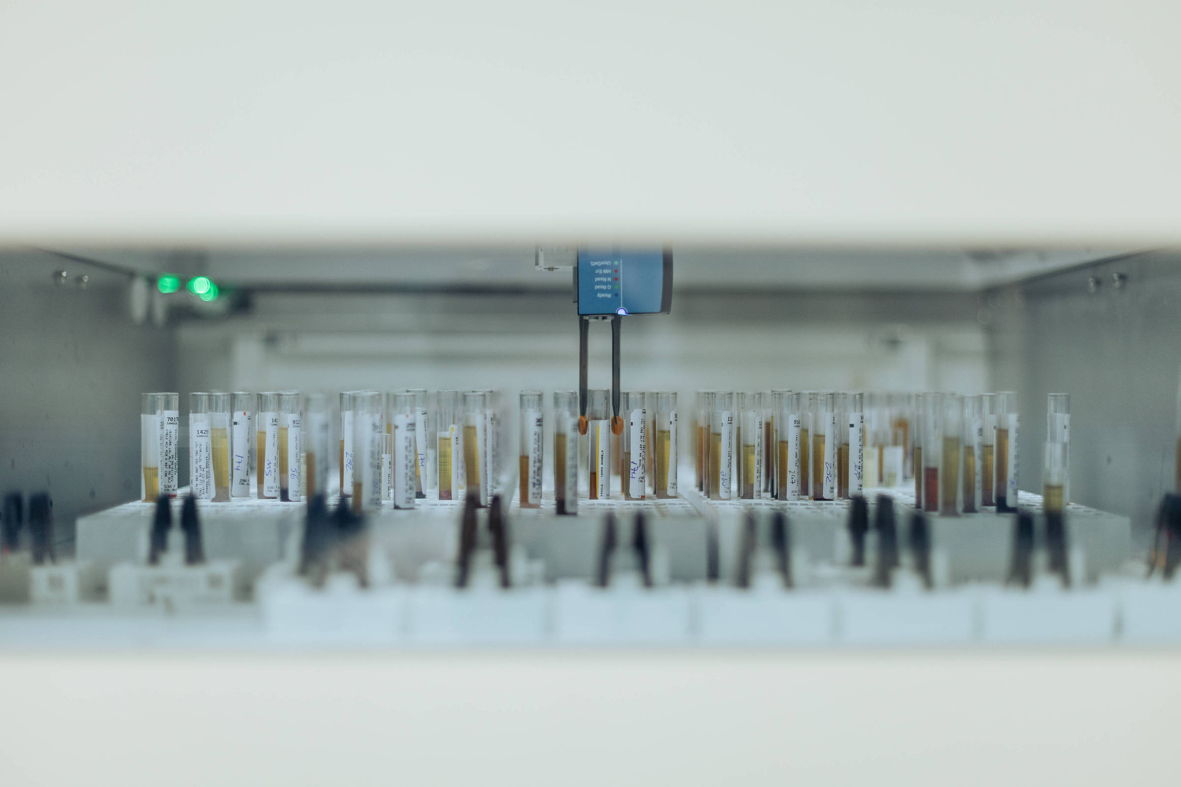 A imagem mostra um equipamento de laboratório com tubos de ensaio dispostos em uma prateleira. Um dispositivo mecânico está posicionado acima dos tubos, aparentemente realizando uma análise ou coleta de amostras. Os tubos contêm líquidos de diferentes cores, e há luzes verdes acesas no equipamento.