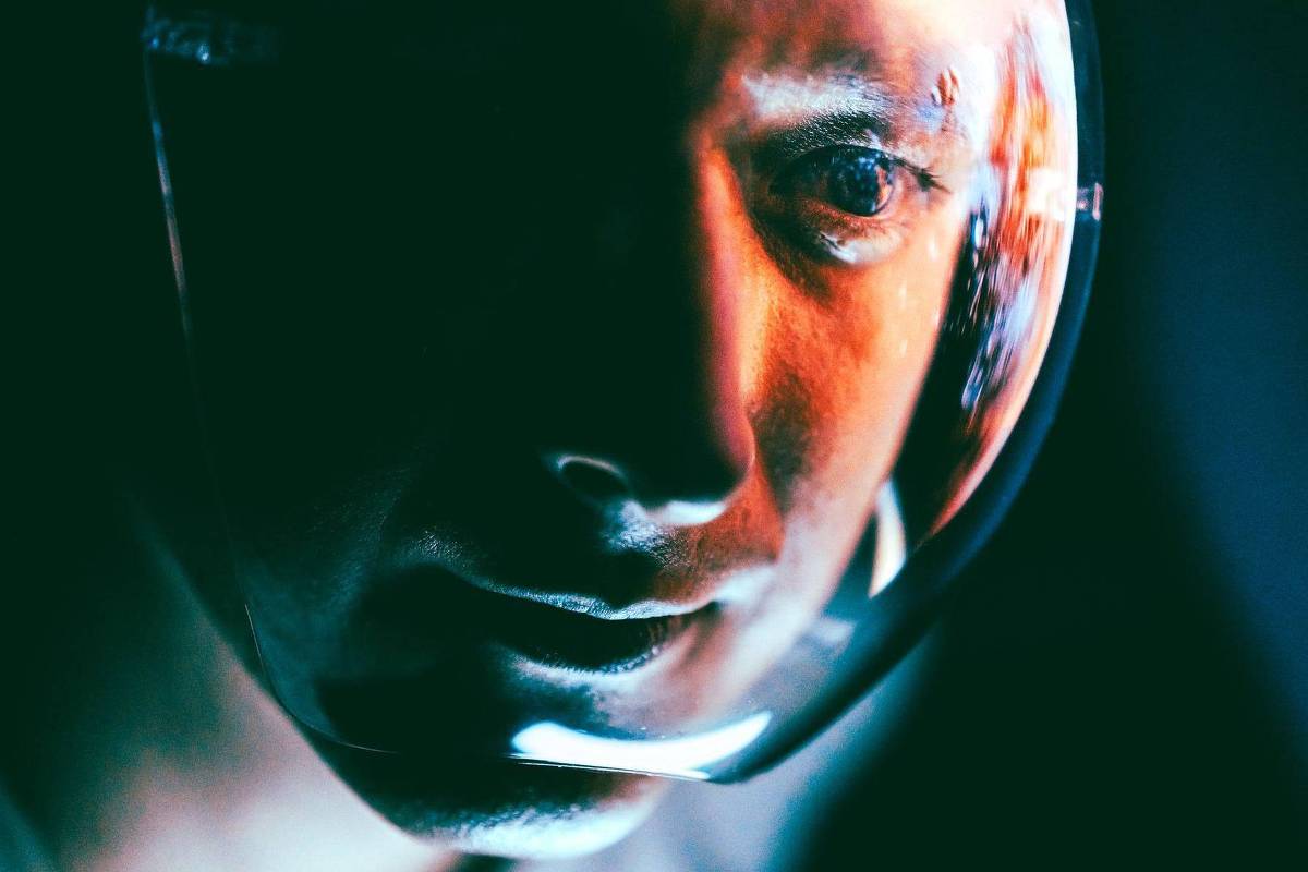 A imagem mostra um close-up do rosto de um astronauta, com um capacete refletivo que destaca a expressão facial. A iluminação é dramática, com tons de azul e laranja, criando um contraste marcante. O fundo é escuro, o que enfatiza a figura do astronauta e o brilho do capacete.