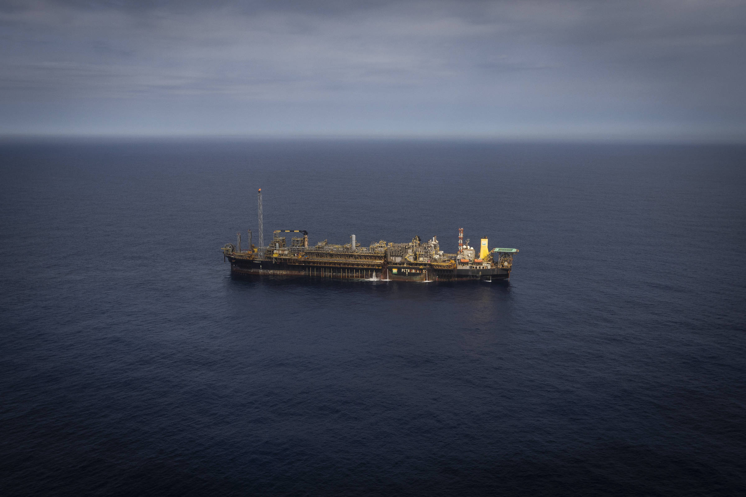 A imagem mostra uma plataforma de petróleo flutuante no meio do oceano. A plataforma é grande e possui várias estruturas e equipamentos visíveis, com um fundo de céu nublado e água calma ao redor.