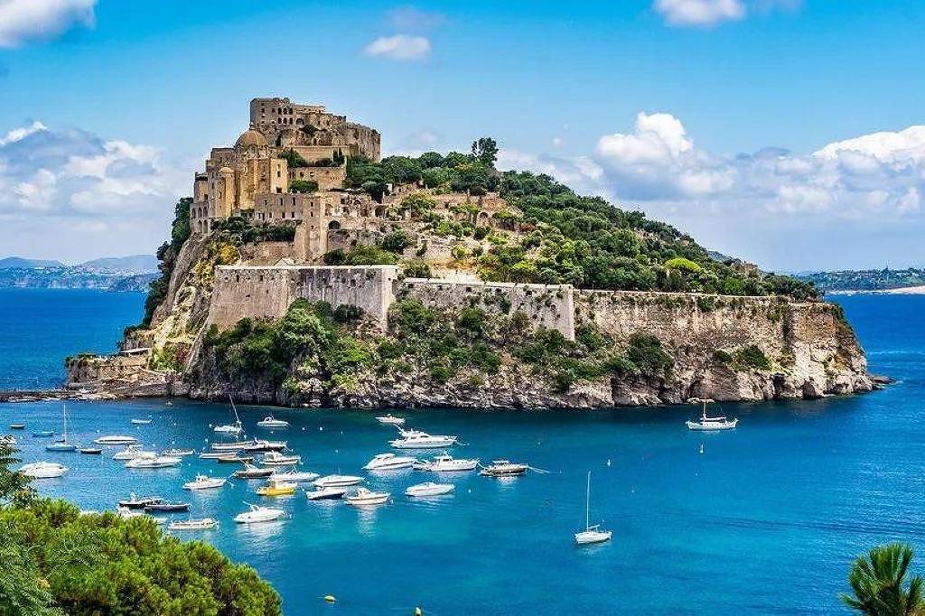 A imagem mostra uma ilha com um castelo no topo, cercada por água azul clara. Vários barcos estão ancorados na baía em frente à ilha. O céu está limpo com algumas nuvens brancas e há vegetação verde ao redor da costa.