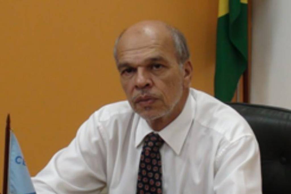 Um homem de cabelos grisalhos está sentado em uma mesa de escritório. Ele usa uma camisa branca e uma gravata com padrões. Ao fundo, há uma bandeira do Brasil e uma bandeira azul. O homem parece sério e está com as mãos apoiadas na mesa de madeira.