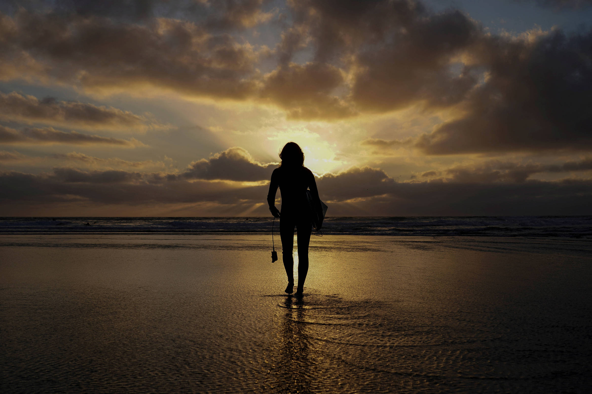 A imagem mostra a silhueta de uma pessoa caminhando na praia durante o pôr do sol. O céu está parcialmente nublado, com nuvens iluminadas por tons de laranja e amarelo. A água do mar reflete a luz do sol, criando um efeito brilhante na superfície. A pessoa está de costas, com uma postura relaxada, e parece estar segurando algo na mão.