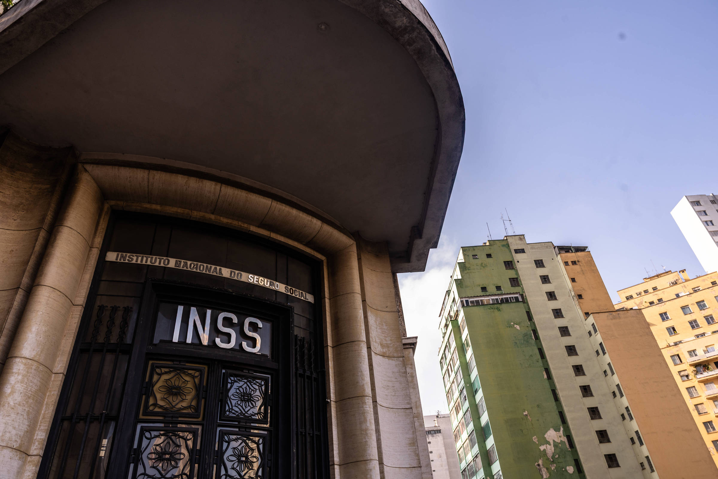 A imagem mostra a fachada de um prédio do INSS (Instituto Nacional do Seguro Social) com uma porta de entrada em destaque. O nome