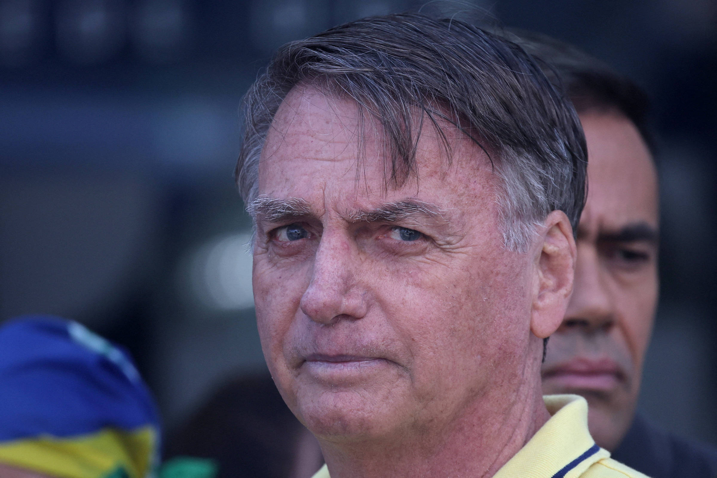 Jair Bolsonaro, um homem com cabelo castanho claro e liso, usando uma camisa amarela. Ele está olhando para a câmera com uma expressão séria. Ao fundo, há outras pessoas desfocadas, e uma bandeira do Brasil é visível em um dos lados da imagem.