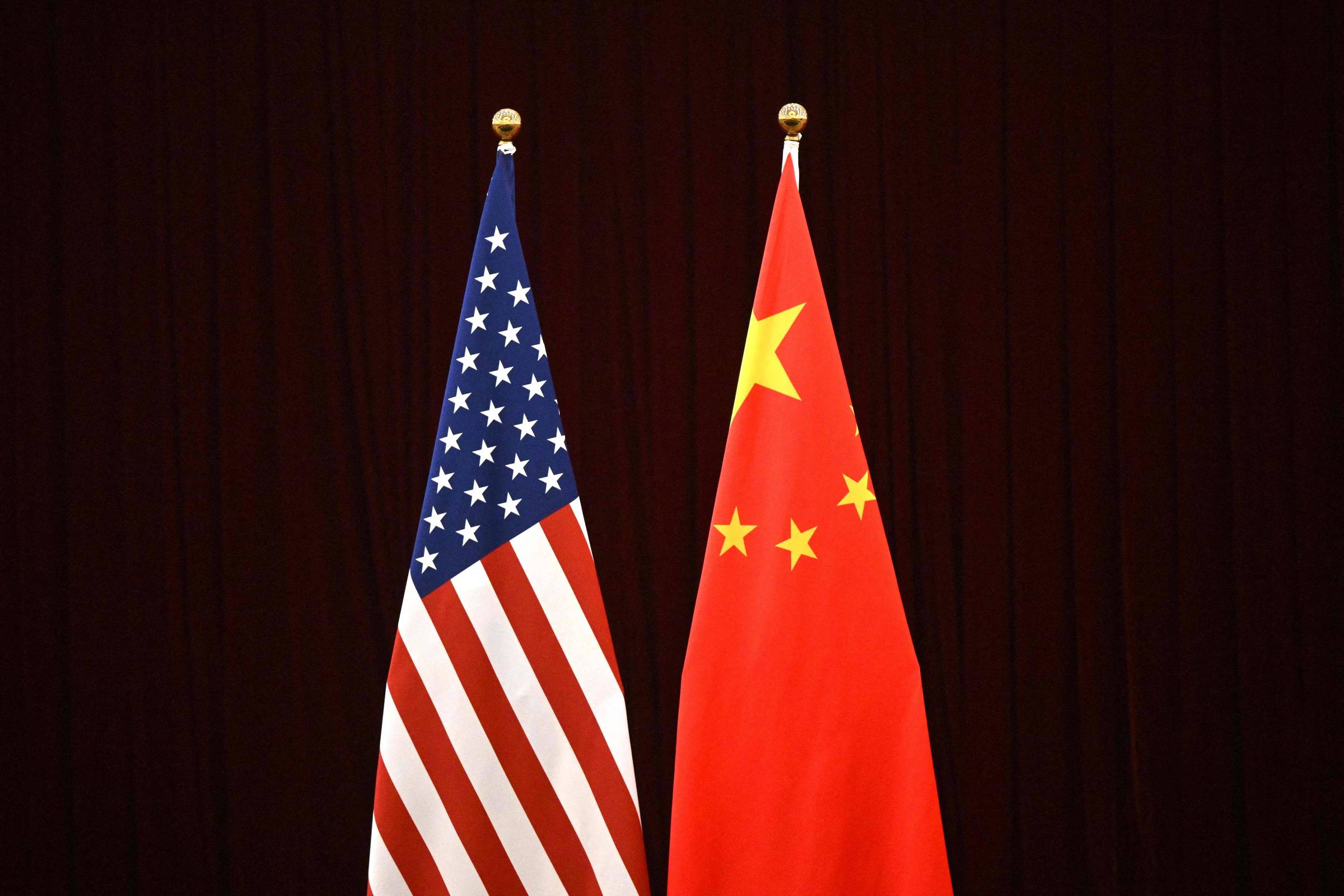 A imagem mostra duas bandeiras: à esquerda, a bandeira dos Estados Unidos, com listras vermelhas e brancas e um campo azul com estrelas brancas; à direita, a bandeira da China, que é vermelha com uma grande estrela amarela e quatro estrelas menores em um canto superior esquerdo. O fundo da imagem é escuro.