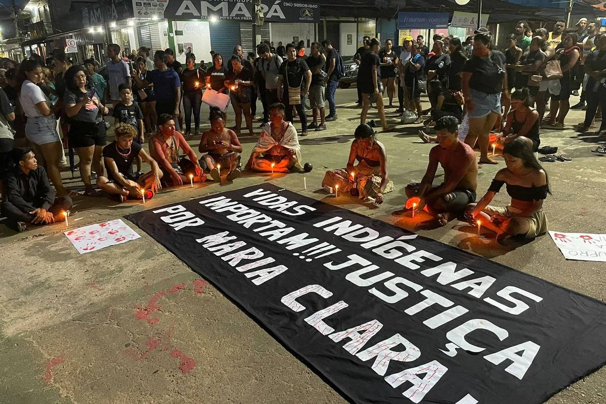 A imagem mostra um grupo de pessoas reunidas em um protesto à noite. Algumas pessoas estão sentadas no chão, segurando velas acesas, enquanto outras estão em pé ao fundo. No centro, há um grande banner preto com letras brancas que diz: