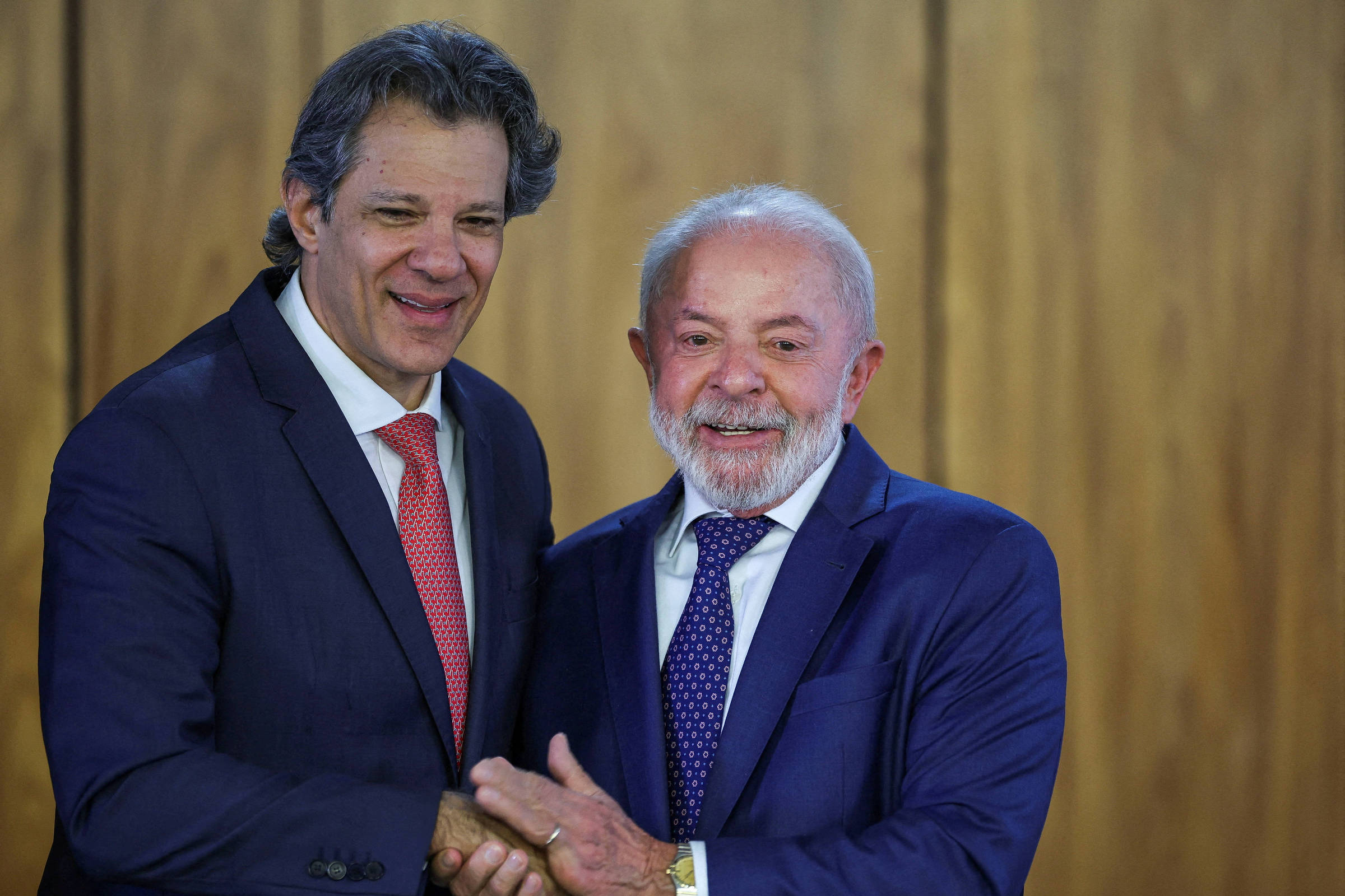 Na imagem, duas pessoas estão se cumprimentando com um aperto de mão. À esquerda, um homem de cabelo grisalho e liso, vestindo um terno escuro com uma gravata vermelha. À direita, um homem mais velho, com cabelo grisalho e barba, usando um terno azul e uma gravata com padrões. O fundo é de madeira clara, sugerindo um ambiente formal.