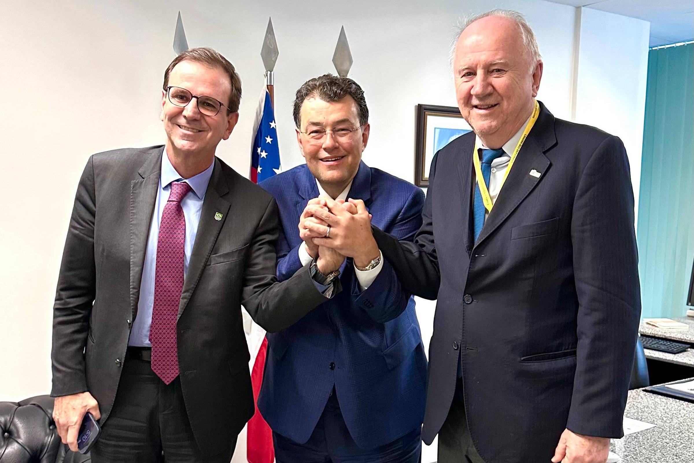 Três homens estão em uma sala, sorrindo e segurando as mãos uns dos outros. O homem do meio está vestido com um terno azul e os outros dois estão usando ternos escuros. Ao fundo, há uma bandeira e um símbolo que parece ser uma lanterna ou uma lança. A atmosfera é de celebração ou camaradagem.