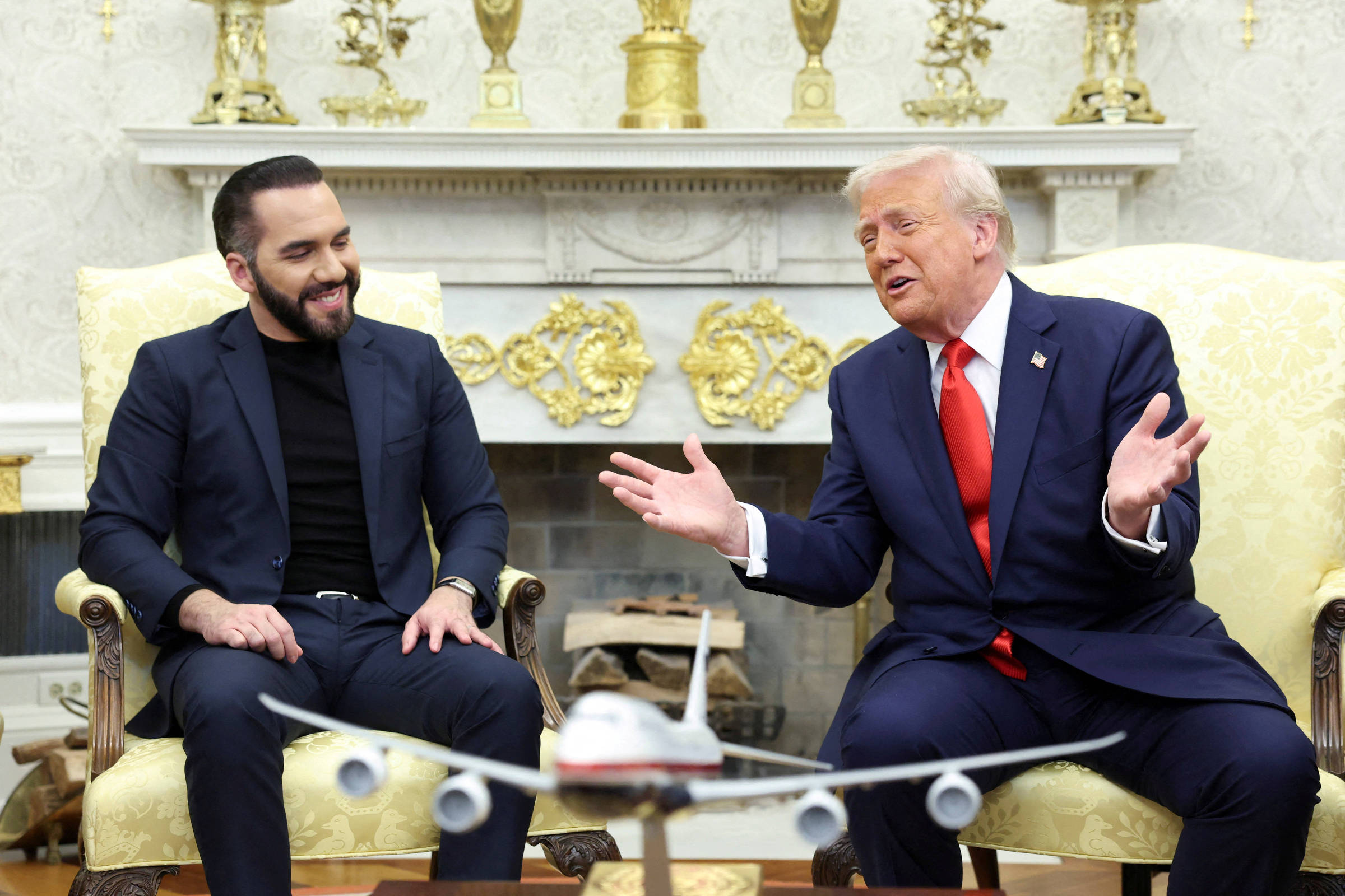 Na imagem, duas pessoas estão sentadas em cadeiras no Salão Oval. À esquerda, um homem com barba e cabelo curto, vestido com uma camisa preta e um blazer, sorri enquanto observa o homem à direita. À direita, um homem com cabelo loiro e terno azul, usando uma gravata vermelha, gesticula com as mãos enquanto fala. Entre eles, há um modelo de avião em uma mesa. O ambiente é decorado com elementos clássicos, incluindo um lareira ao fundo e ornamentos dourados.