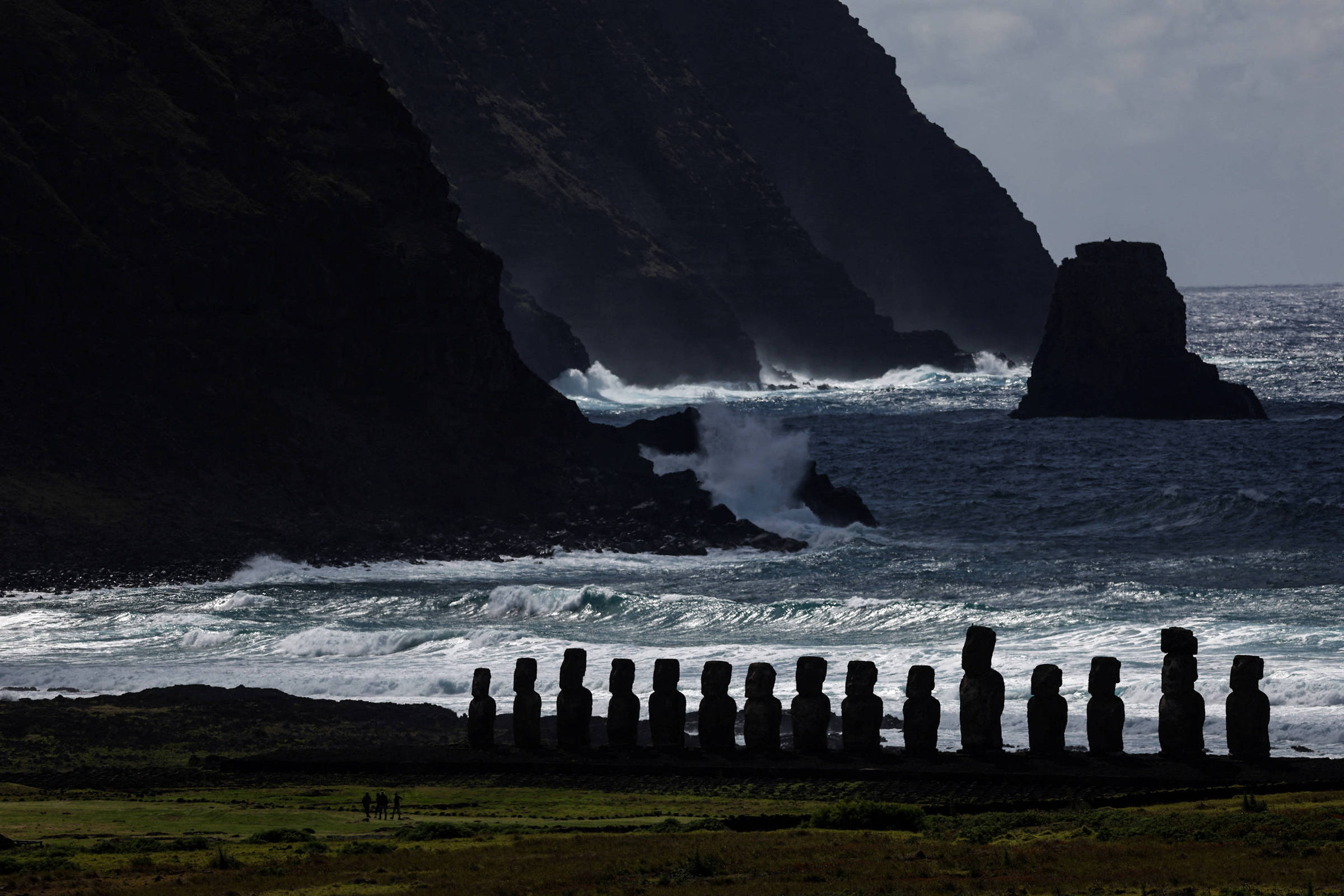 A imagem mostra uma linha de estátuas Moai, típicas da Ilha de Páscoa, em primeiro plano, com o mar agitado ao fundo. As estátuas estão posicionadas em uma área costeira, com montanhas e rochas visíveis nas laterais. O céu está nublado, e as ondas do mar quebram contra a costa, criando um contraste entre a luz e a sombra.