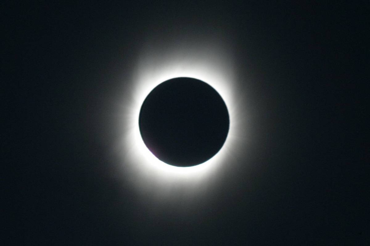 A imagem mostra um eclipse solar total, onde a Lua está cobrindo completamente o Sol. Ao redor da Lua, há uma coroa de luz solar visível, criando um efeito brilhante em contraste com o fundo escuro do céu.