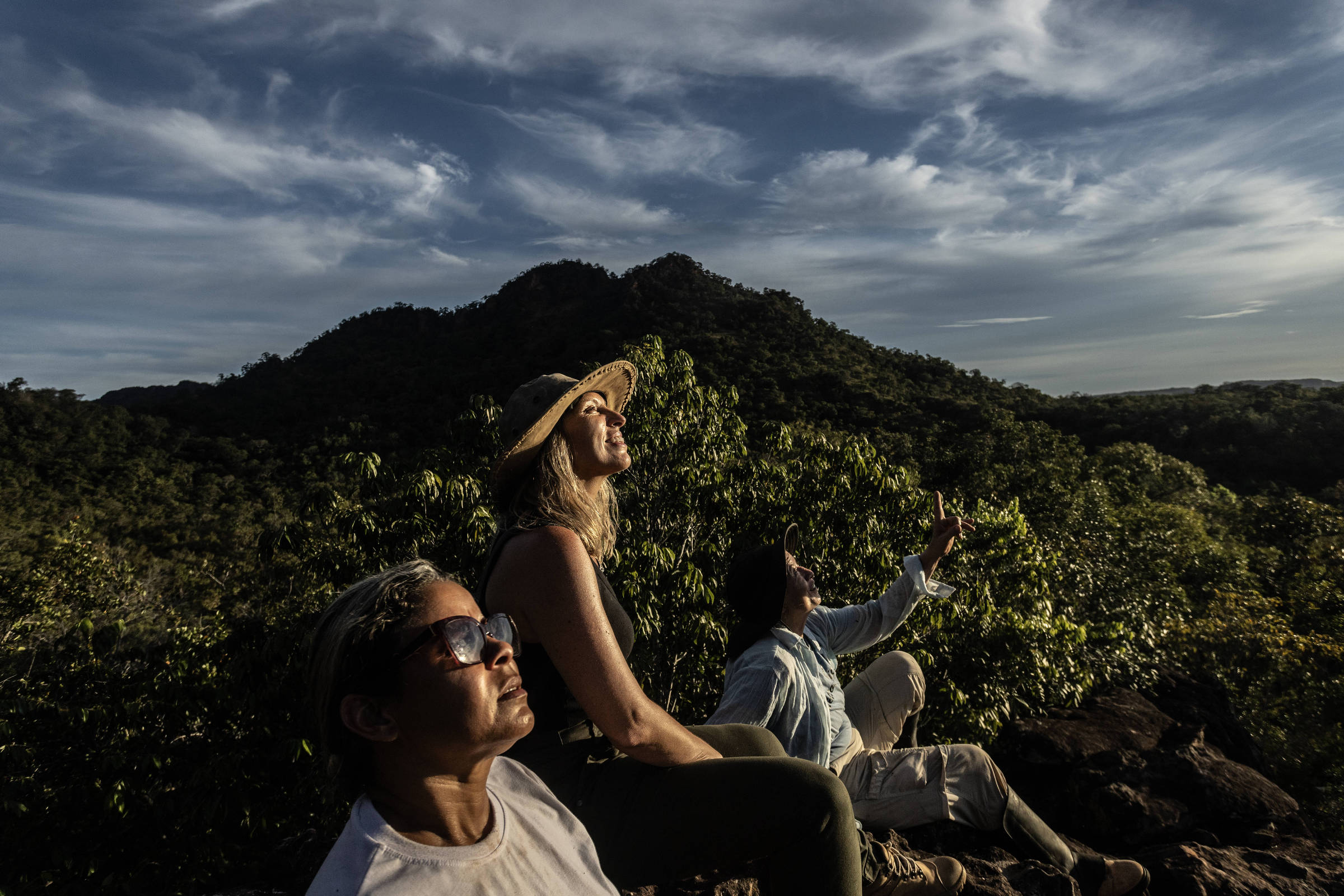 Três pessoas estão sentadas em uma rocha, olhando para o céu. A mulher do meio usa um chapéu e parece estar contemplando algo. As outras duas mulheres estão em posições diferentes, uma com óculos escuros e a outra com uma camisa clara. Ao fundo, há uma paisagem montanhosa coberta de vegetação e um céu com nuvens.
