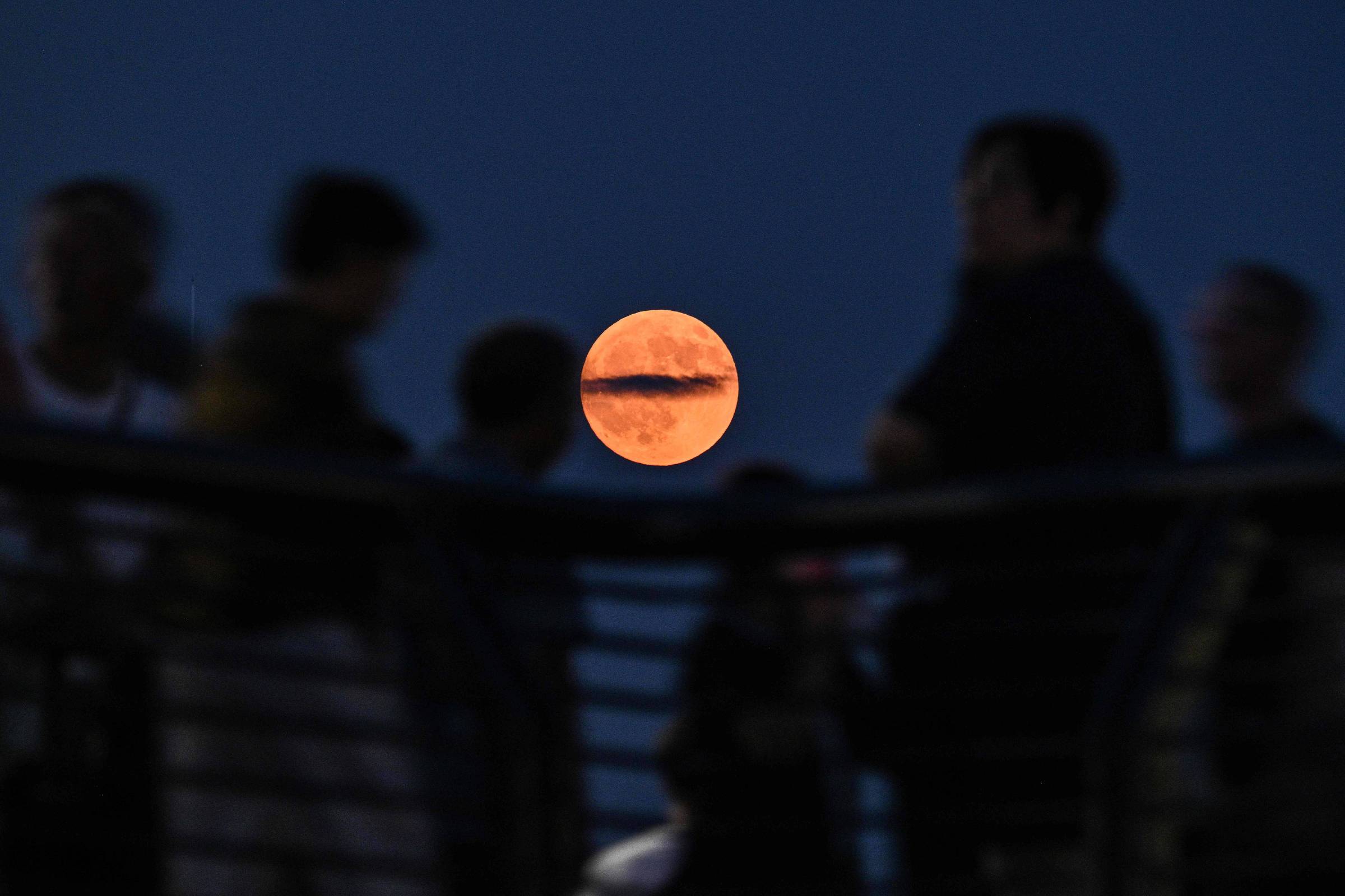 A imagem mostra uma lua cheia de cor laranja brilhante posicionada no centro, com várias silhuetas de pessoas em primeiro plano, que parecem estar observando a lua. O fundo é um céu azul escuro, criando um contraste entre a lua iluminada e as figuras escuras das pessoas.