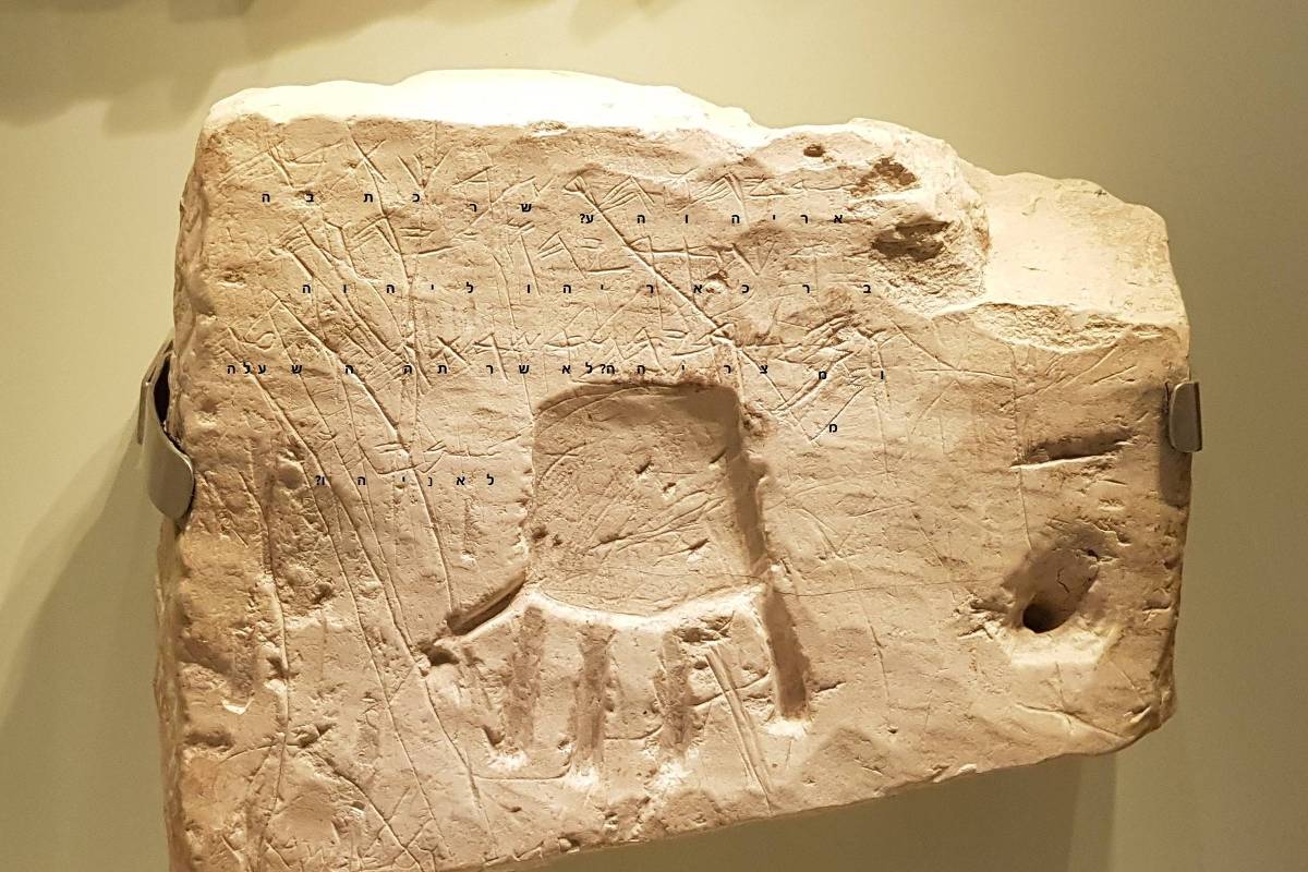A imagem mostra uma placa de pedra antiga com inscrições e desenhos. A placa é de cor clara e apresenta marcas e símbolos gravados em sua superfície, incluindo um desenho que se assemelha a uma figura com braços e um corpo. As inscrições estão dispostas em linhas, sugerindo um texto ou uma forma de comunicação escrita.