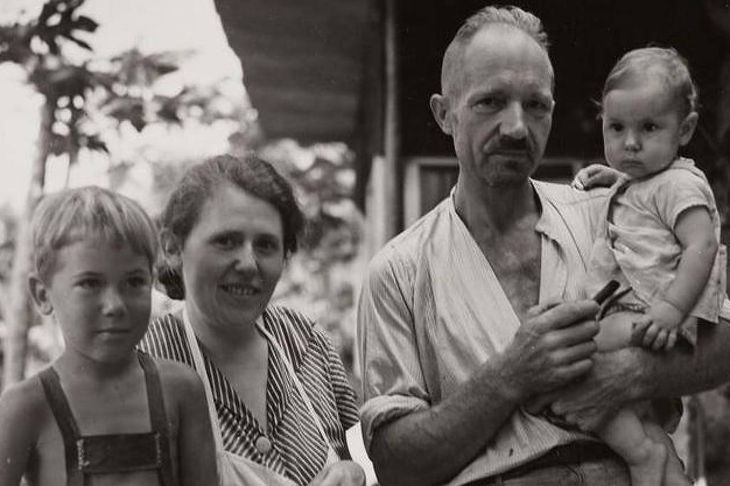 A imagem mostra uma família em um ambiente externo. Há um homem segurando uma criança pequena no colo, uma mulher ao lado dele e um menino à esquerda. O homem tem cabelo curto e usa uma camisa clara, enquanto a mulher tem cabelo preso e veste uma blusa listrada. O menino à esquerda usa um macacão. O fundo é composto por árvores e uma estrutura que parece ser uma casa.