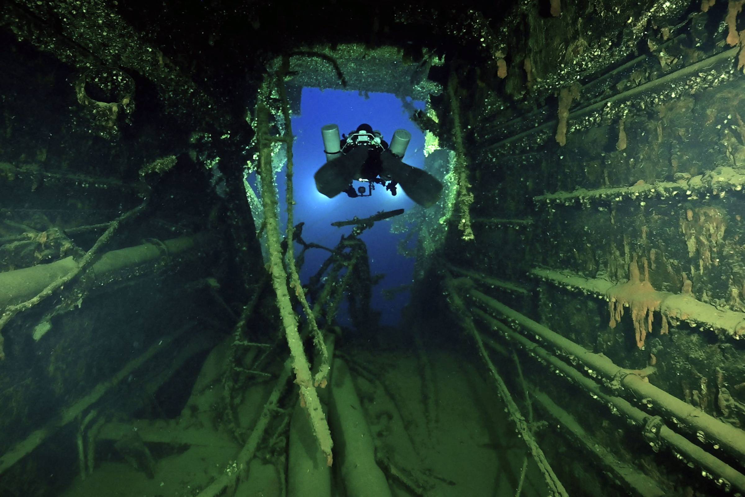 A imagem mostra o interior de um navio afundado, com estruturas metálicas visíveis e cobertas por algas e sedimentos. Um mergulhador está posicionado em um espaço amplo, iluminado por uma luz azul que entra por uma abertura na parte superior. O ambiente é escuro e misterioso, com a presença de detritos e vegetação marinha ao redor.