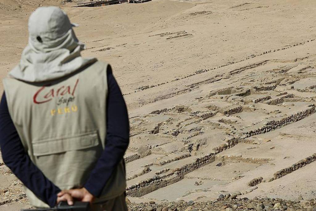 Um arqueólogo, vestido com um colete claro e um chapéu, está de costas, observando um sítio arqueológico em uma área desértica. No chão, há formações de pedras dispostas em linhas retas, que parecem ser vestígios de estruturas antigas. O ambiente é árido e sem vegetação, com algumas marcas no solo visíveis ao fundo.