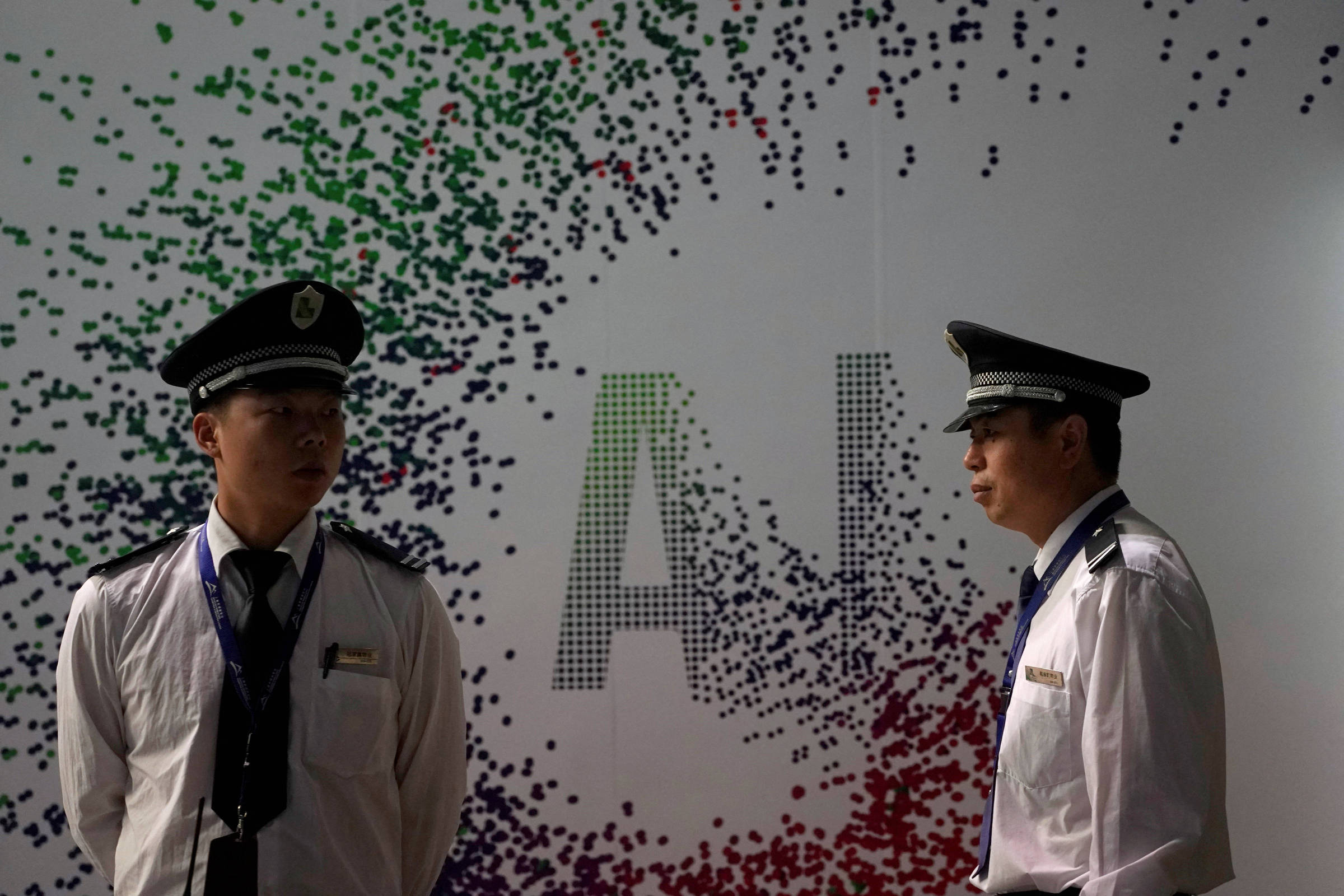 Dois homens em uniformes de segurança estão em pé, um de frente para o outro, em um ambiente que parece ser um evento relacionado à inteligência artificial. Ao fundo, há uma parede decorada com um padrão de pontos coloridos, e a sigla