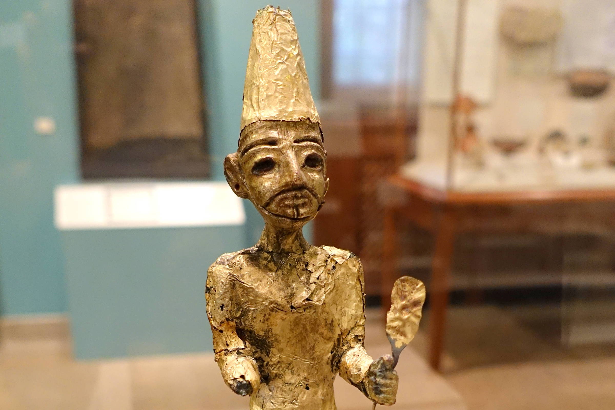 A imagem mostra uma estátua de um personagem humano sentado, feita de metal. O personagem tem um rosto expressivo, com um nariz proeminente e olhos grandes. Ele usa um manto longo e possui um chapéu ou coroa alta. Em uma das mãos, ele segura um objeto, possivelmente um cetro ou bastão. A estátua está sobre uma base de madeira e é exibida em um ambiente de museu, com outros objetos visíveis ao fundo.