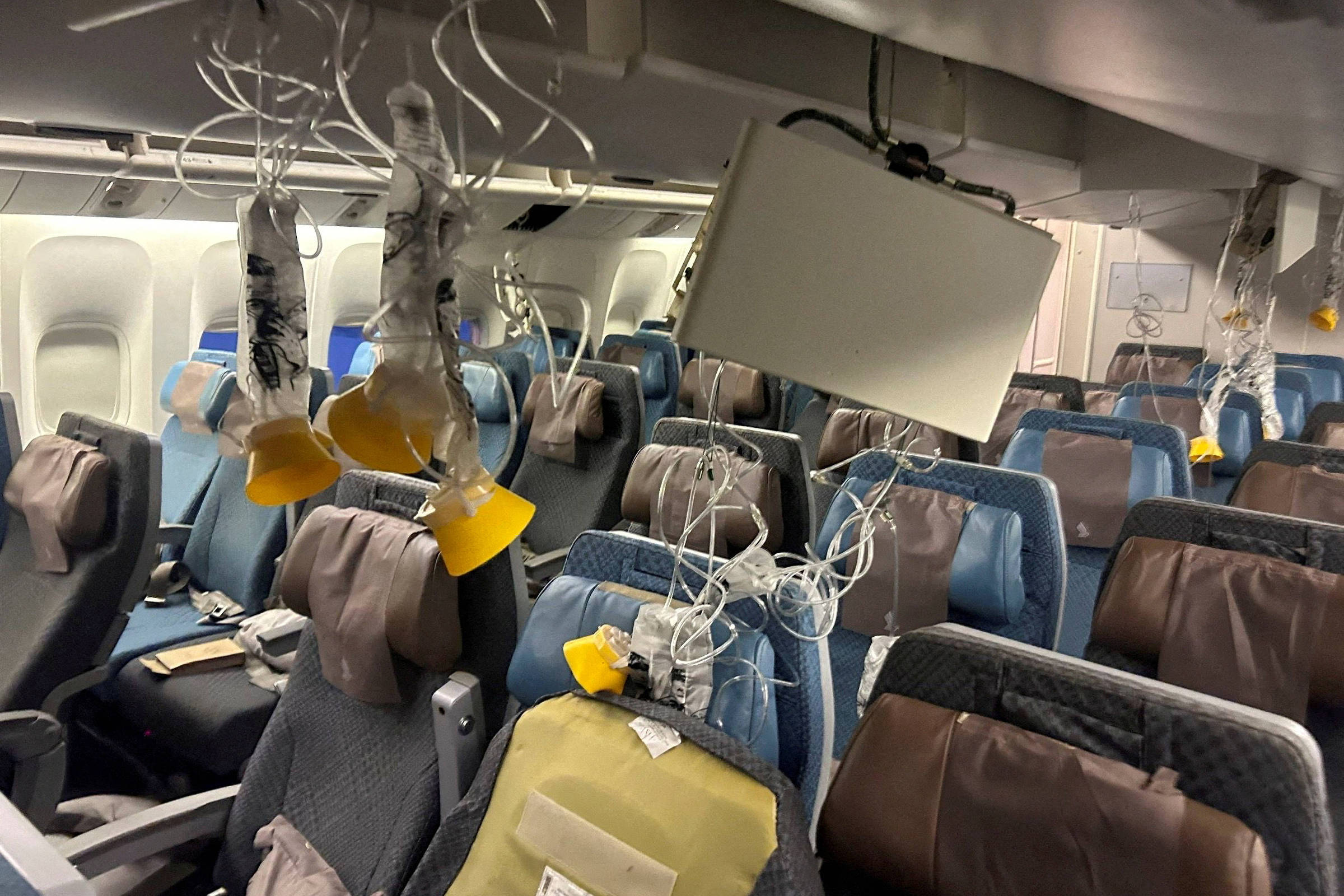 A imagem mostra o interior de uma cabine de avião com várias máscaras de oxigênio penduradas. As máscaras estão conectadas a tubos e algumas estão caídas, enquanto outras estão penduradas no teto. As poltronas estão em um tom de azul e cinza, com capas de almofadas em marrom. O ambiente parece desordenado, sugerindo uma situação de emergência.
