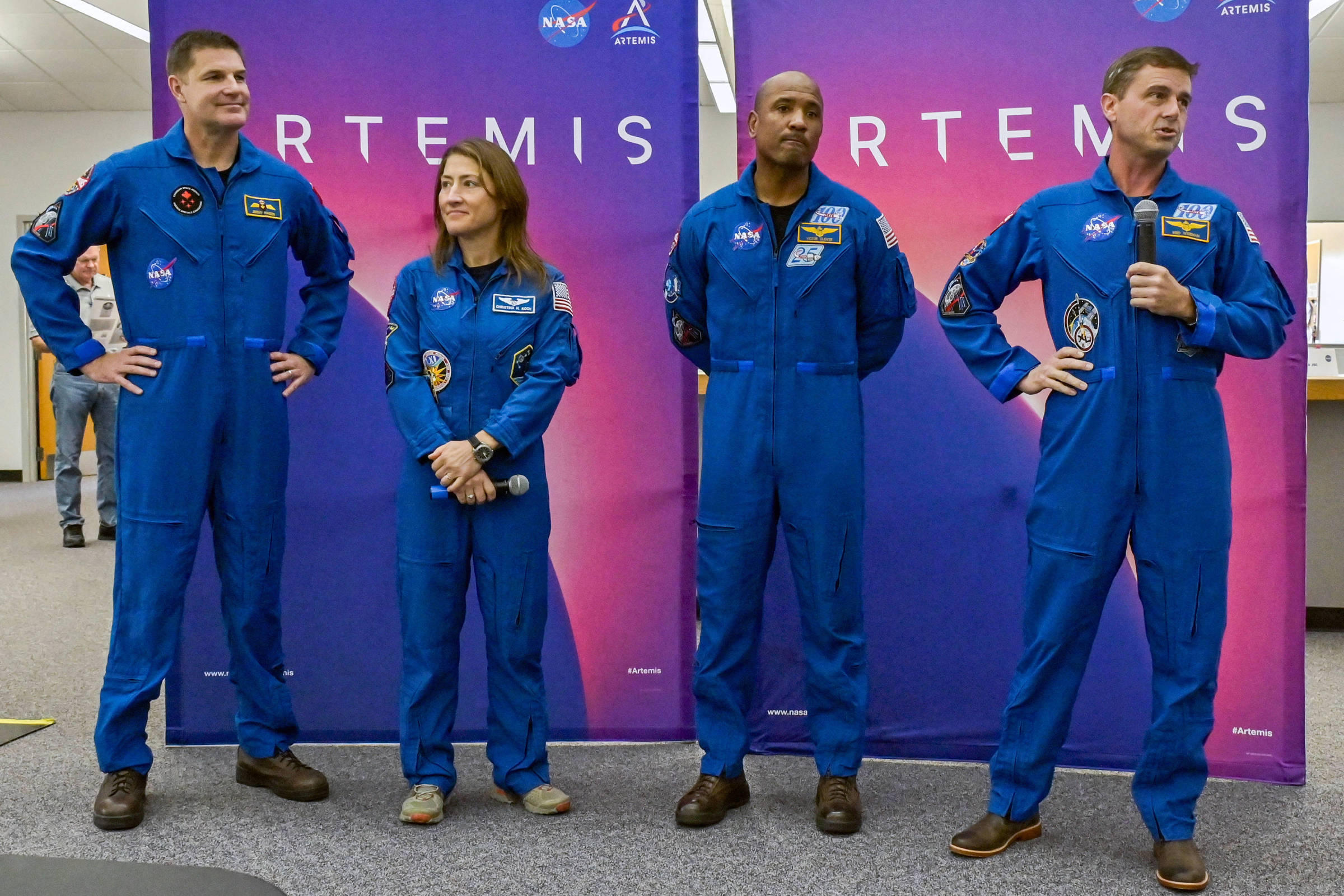 Quatro astronautas vestindo macacões azuis da NASA posam em frente a painéis com o logo da missão Artemis. Dois homens estão nas extremidades, uma mulher e um homem no centro, todos em ambiente interno.