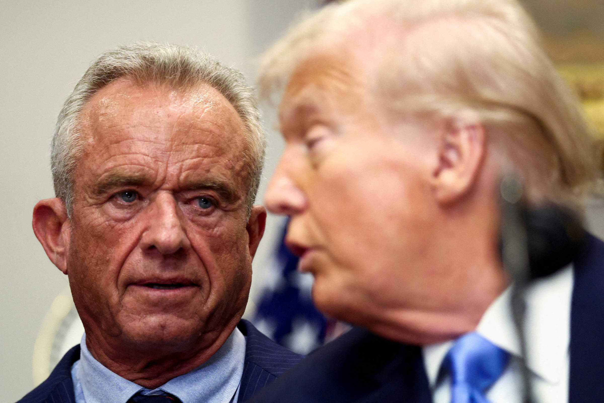 Robert F. Kennedy Jr. olha atentamente para Donald Trump durante evento oficial. Trump está em primeiro plano, parcialmente desfocado, com terno escuro e gravata azul.