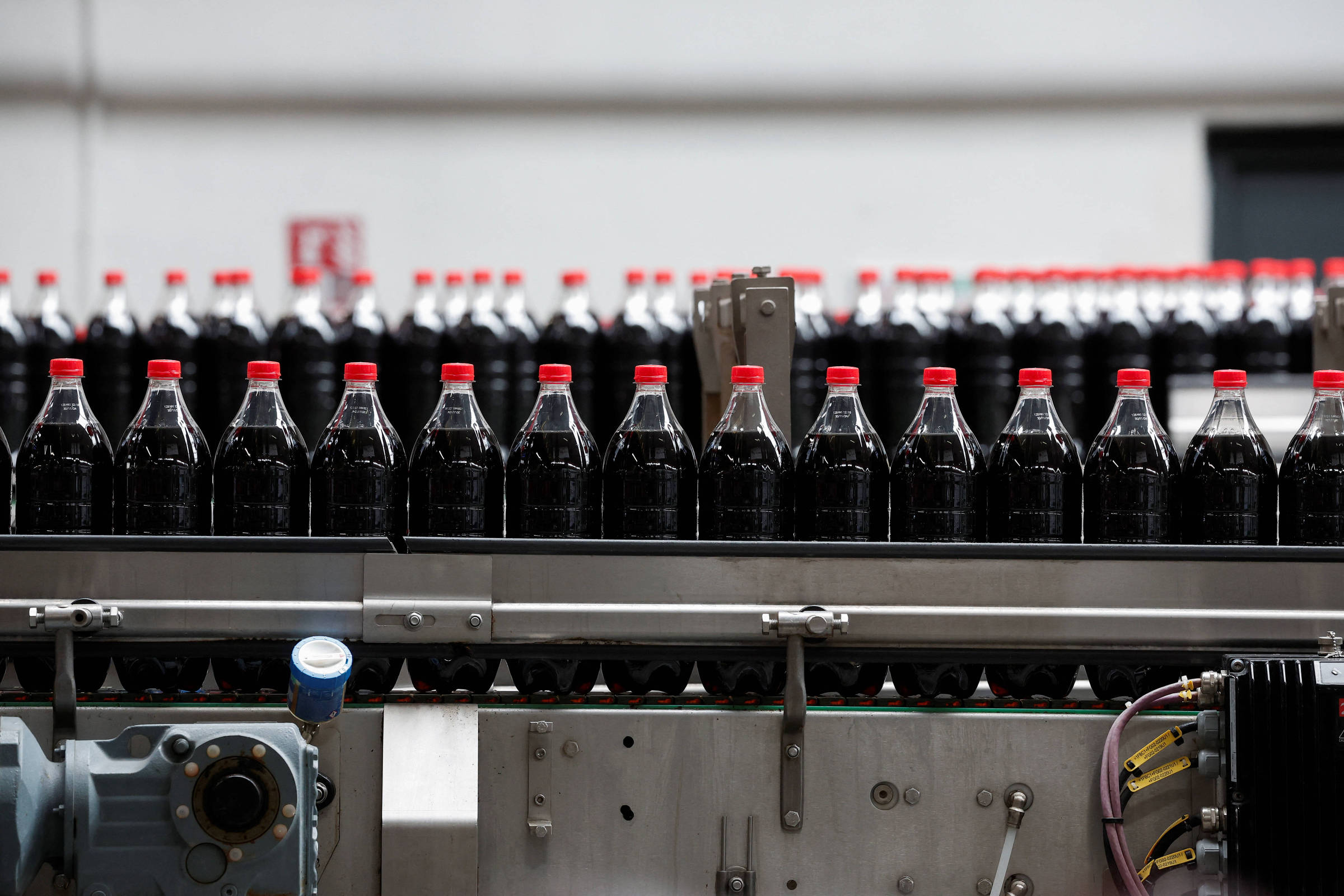 Diversas garrafas plásticas de refrigerante com tampas vermelhas alinhadas em uma esteira de produção industrial. Equipamentos e componentes da máquina são visíveis na parte inferior da imagem.