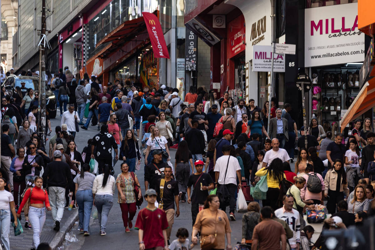 A imagem mostra uma rua comercial movimentada, cheia de pessoas caminhando. Há lojas com fachadas visíveis, algumas com letreiros e produtos expostos. O ambiente é urbano, com uma grande quantidade de pedestres, incluindo pessoas de diferentes idades e estilos. A cena transmite uma sensação de atividade e comércio.