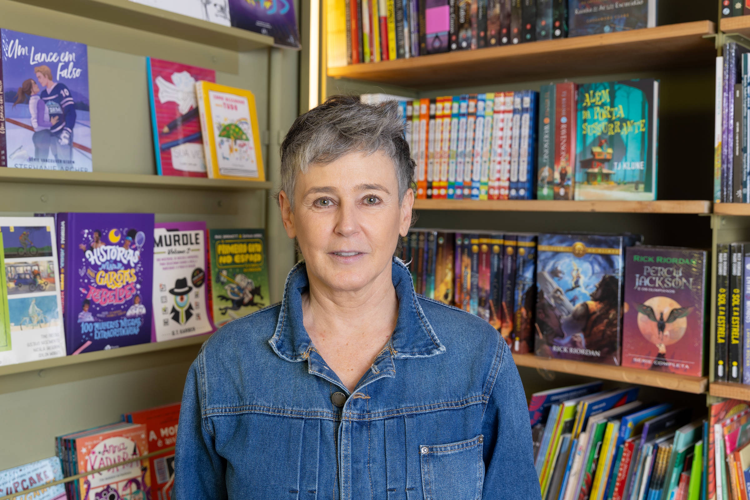 Uma mulher de cabelo curto e grisalho está em pé em uma livraria, com prateleiras de livros ao fundo. Ela usa uma jaqueta jeans e parece estar olhando para a câmera. As prateleiras estão cheias de livros de diferentes cores e tamanhos, incluindo capas de livros infantis e de ficção.