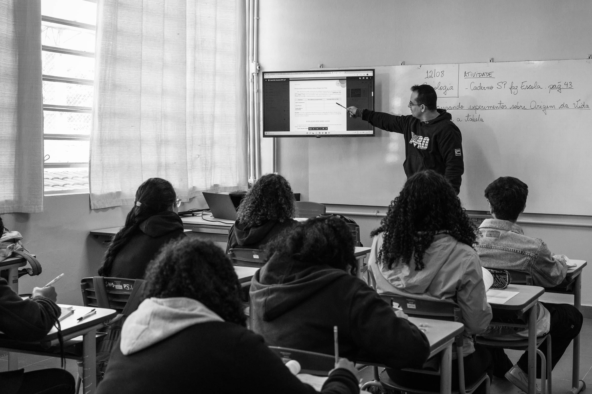 A imagem mostra uma sala de aula com um professor em pé, apontando para uma tela de projeção. O professor está vestido com uma camiseta preta e está de frente para os alunos, que estão sentados em carteiras. A sala tem janelas com cortinas claras e uma lousa branca ao fundo, onde há anotações. Os alunos estão concentrados, alguns escrevendo em cadernos e outros olhando para a tela.