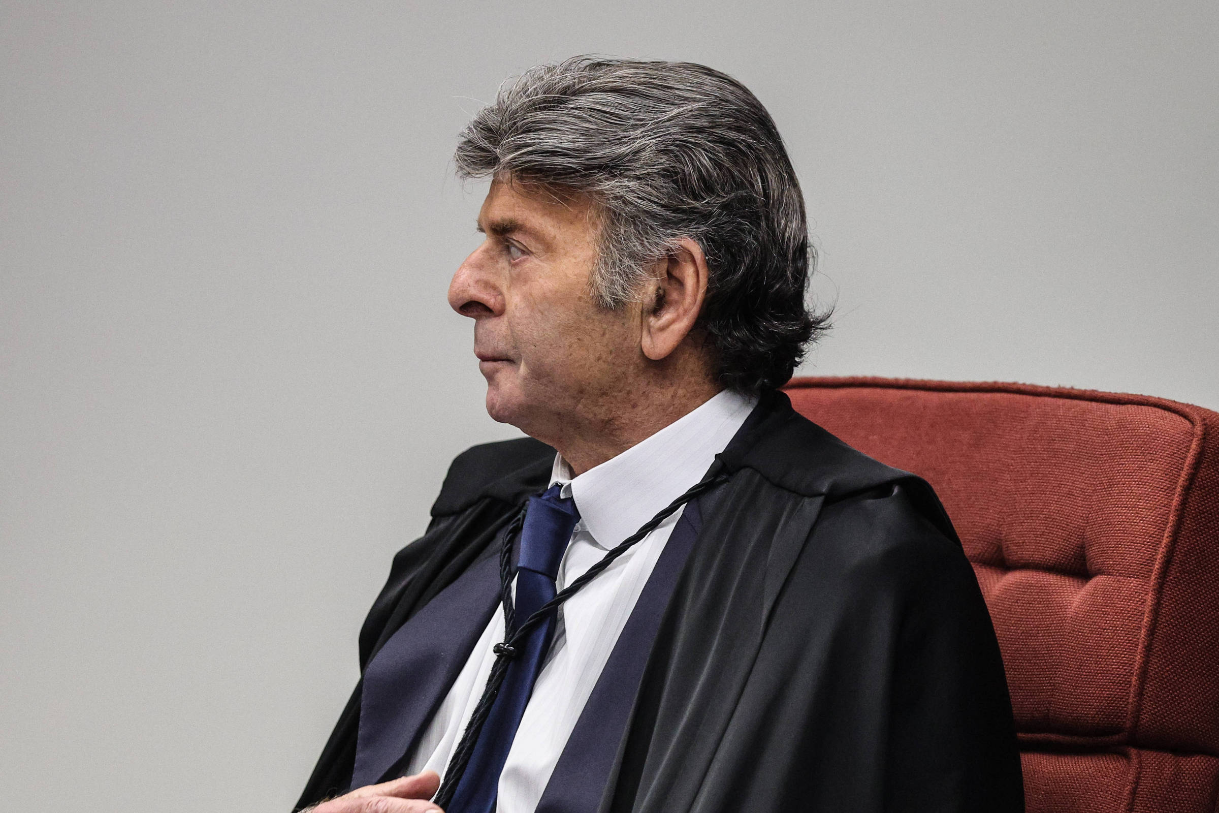 Um homem com cabelo grisalho e liso, usando uma toga preta e uma gravata azul, está sentado em uma cadeira vermelha. Ele está de perfil, olhando para o lado, com uma expressão séria. O fundo é neutro e claro, destacando a figura do homem.