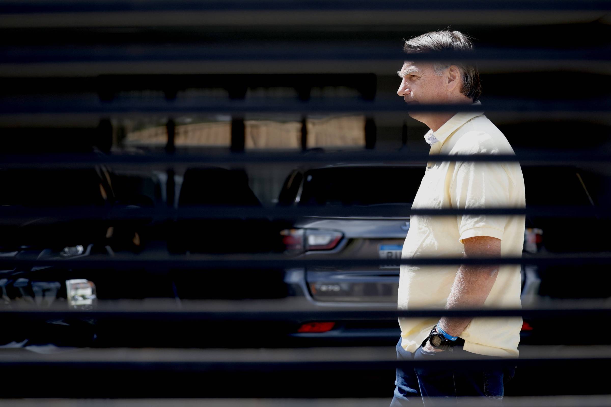 A imagem mostra um homem caminhando em um estacionamento, visível através de uma grade. Ele está usando uma camisa amarela e calças escuras. Ao fundo, há carros estacionados, com um foco em um veículo cinza. A iluminação sugere que é um dia ensolarado.