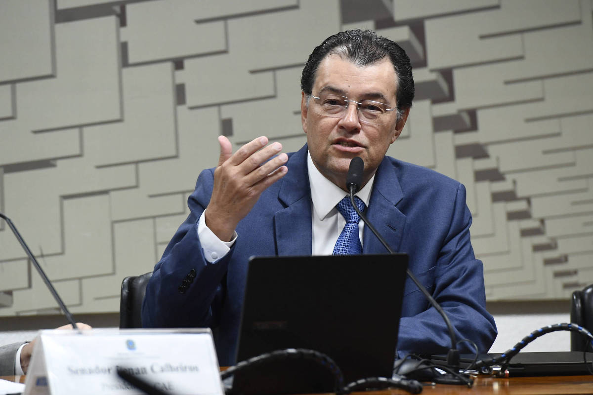 Um homem de terno azul e gravata, com cabelo escuro e óculos, está falando em um microfone durante uma audiência. Ele gesticula com a mão direita enquanto se dirige a um público. Na mesa à sua frente, há um laptop e um documento com o nome