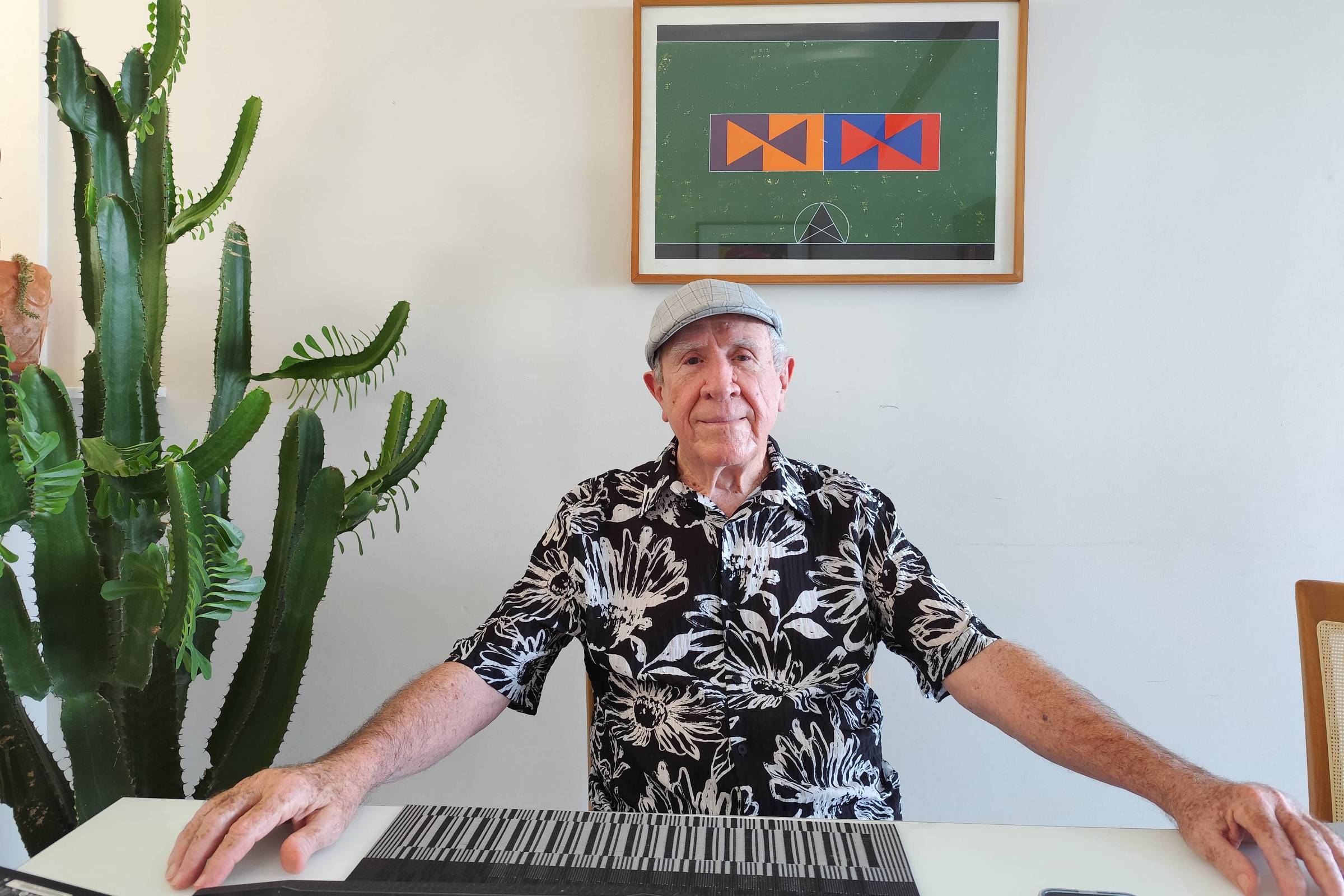 Homem idoso usando boné cinza e camisa preta com estampa branca está sentado à mesa branca. À esquerda, há um grande cacto verde e, ao fundo, uma parede branca com quadro colorido emoldurado.