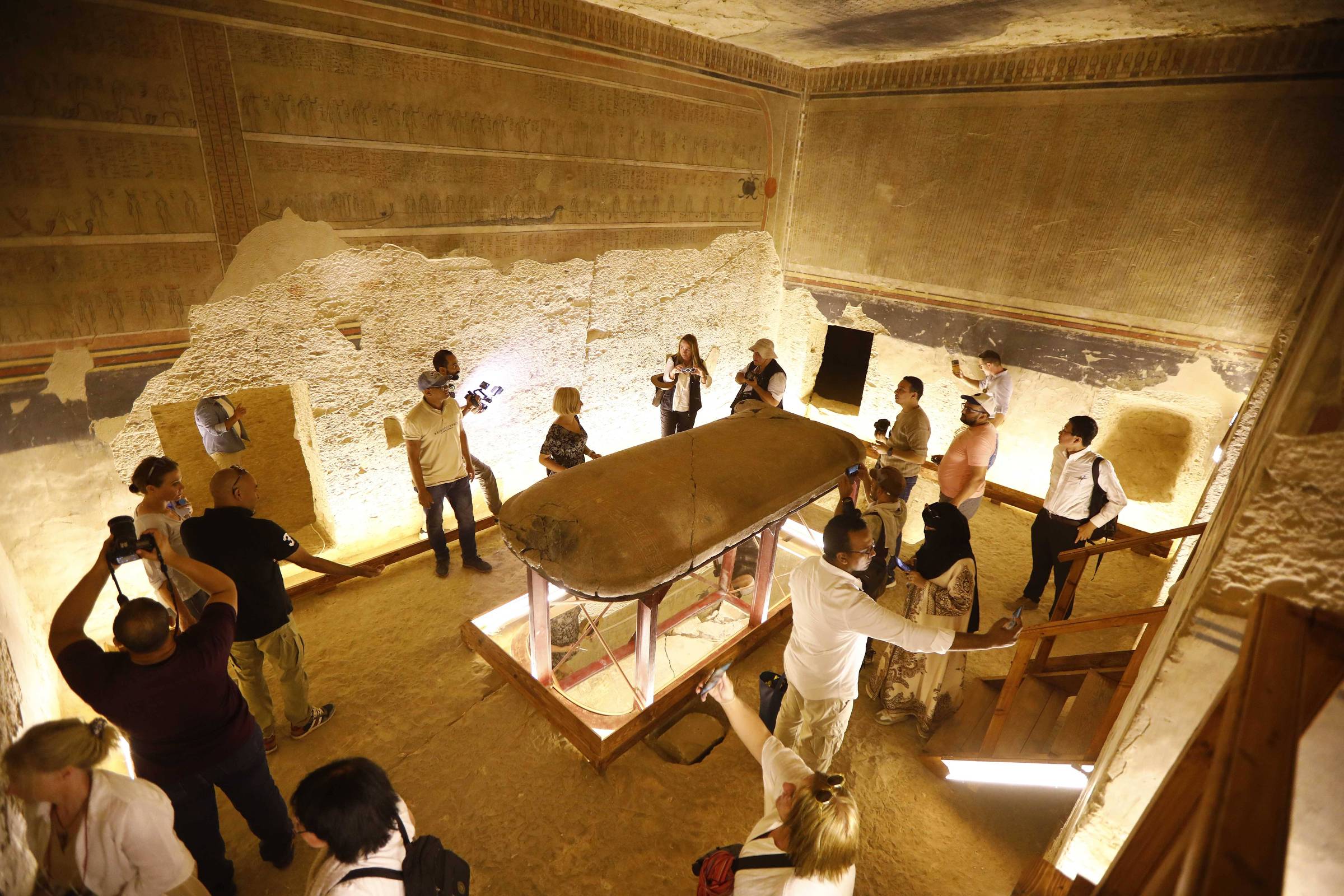 Grupo de pessoas observa e fotografa sarcófago exposto em sala com paredes antigas e iluminação indireta. Ambiente interno com estrutura histórica visível.