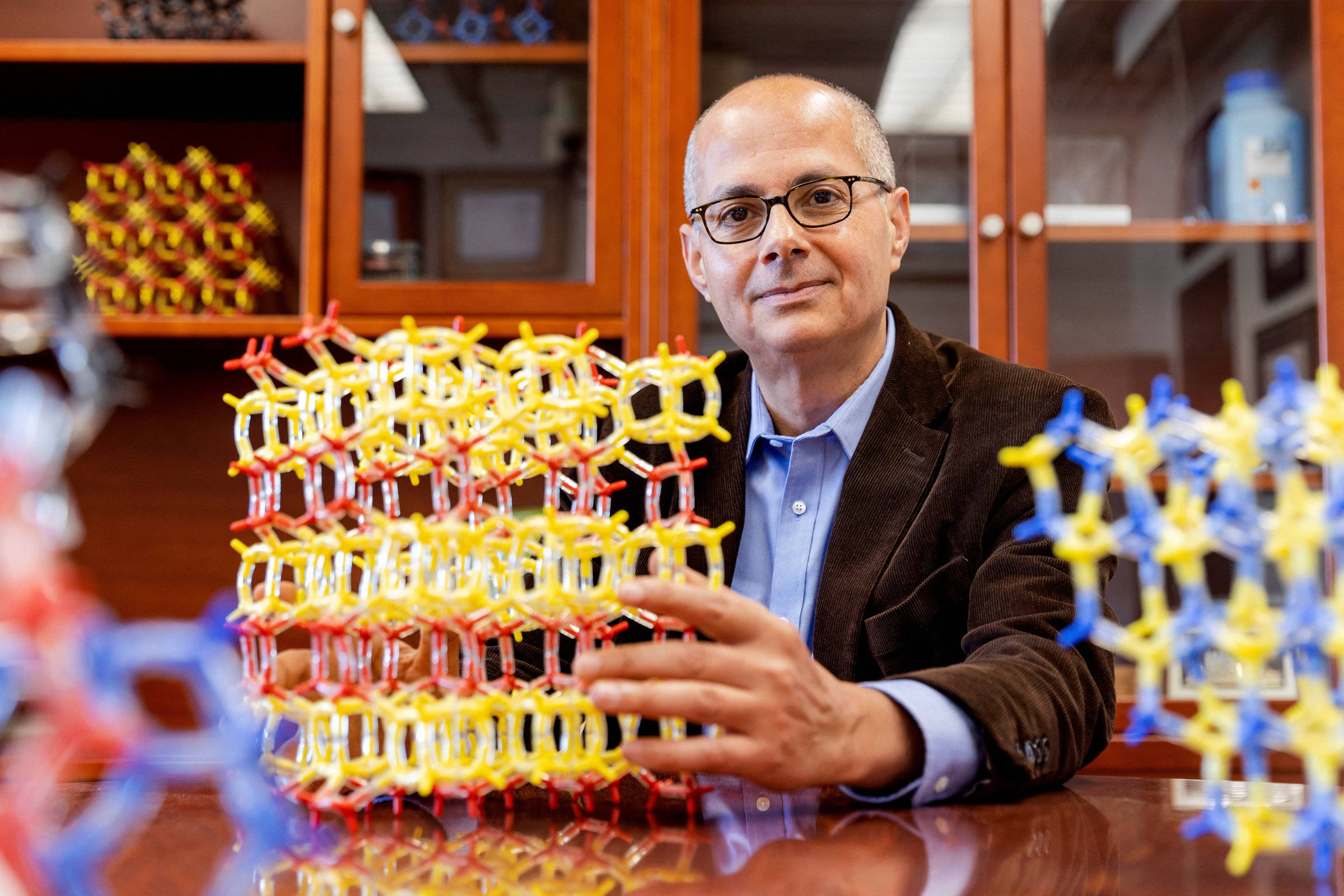 Homem de óculos e blazer marrom segura estrutura molecular colorida em mesa de laboratório. Estantes com livros e objetos científicos ao fundo.