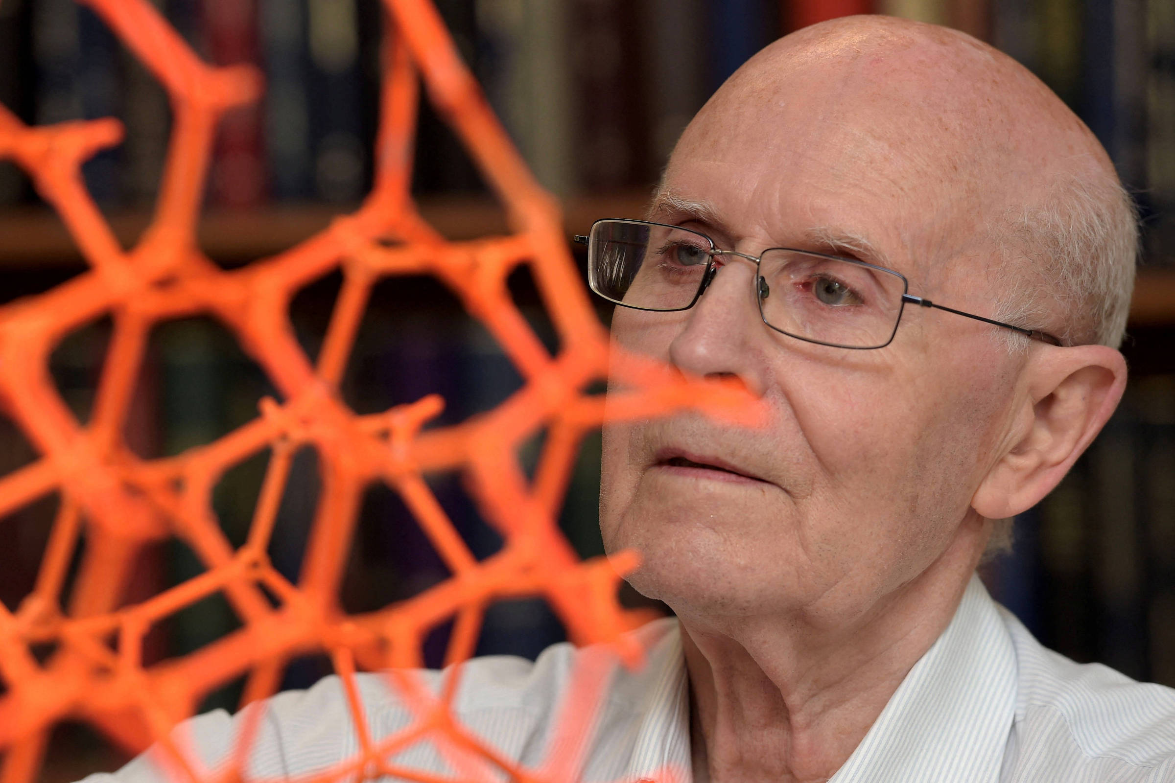Homem idoso de óculos e camisa branca observa atentamente uma estrutura geométrica laranja translúcida. Ao fundo, estantes com livros em ambiente interno.