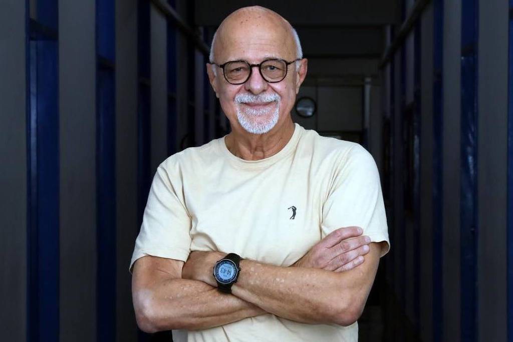 Homem idoso de cabelos brancos e careca, usando óculos e camiseta clara, está com os braços cruzados em corredor interno com paredes azuis.